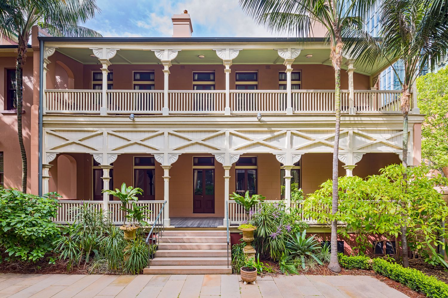 Historic building entrance with a symmetrical double verandah, ornate columns, central stairs and a lush garden of palms and shrubs.