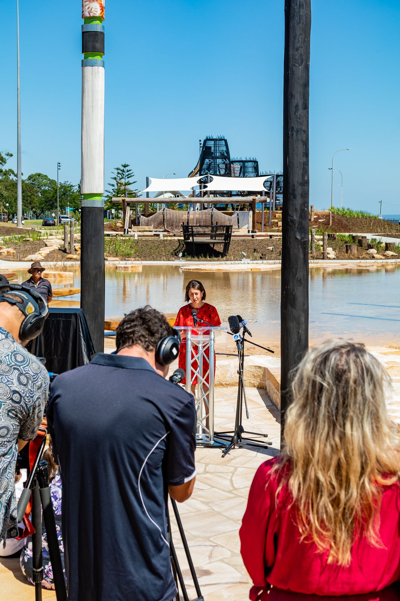 Speaker at a podium by a waterfront plaza while media crew with headphones record, with painted poles and play structure behind.