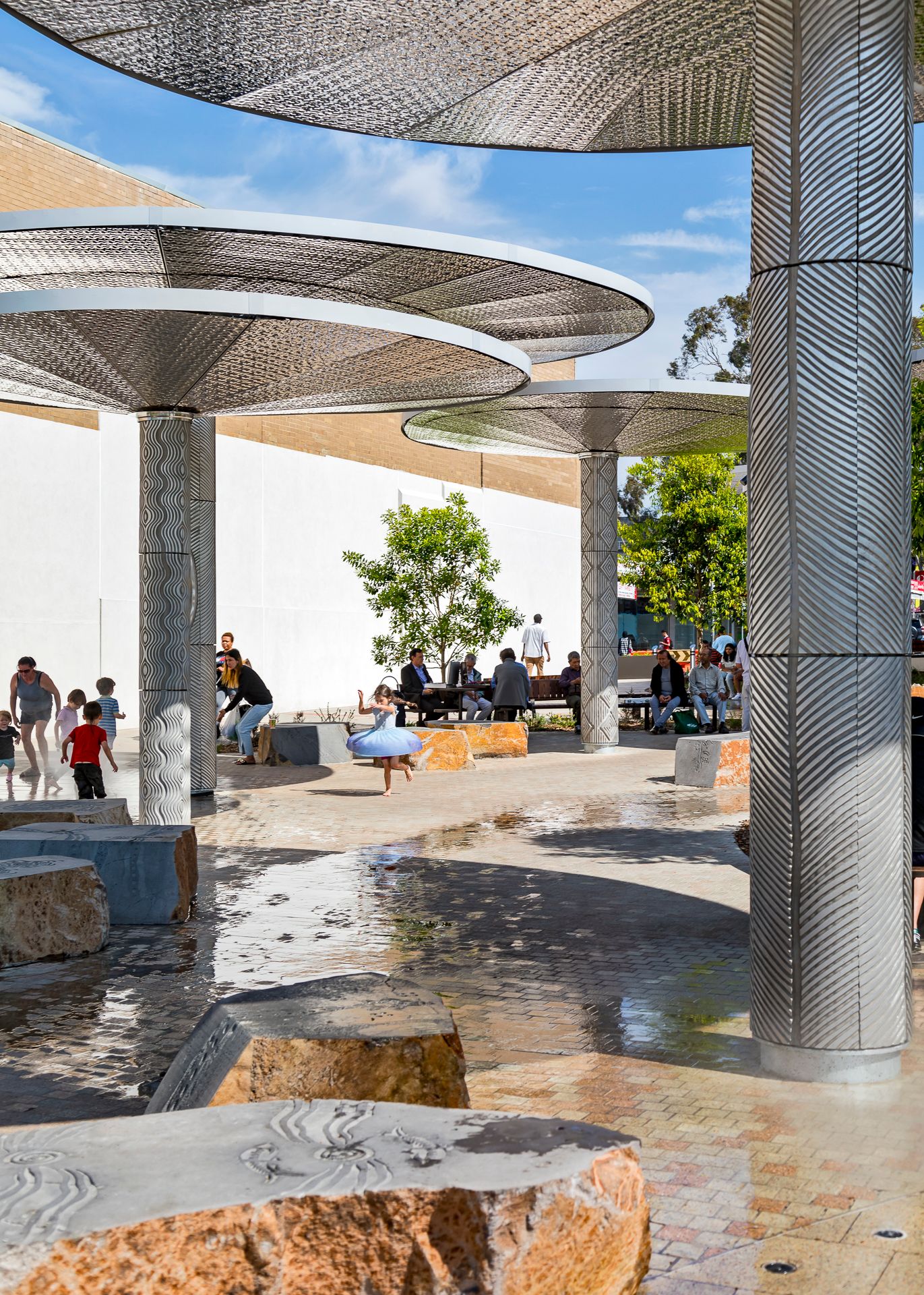 Children play on wet paving near carved sandstone boulders beneath perforated metal canopies in a sunlit public square.