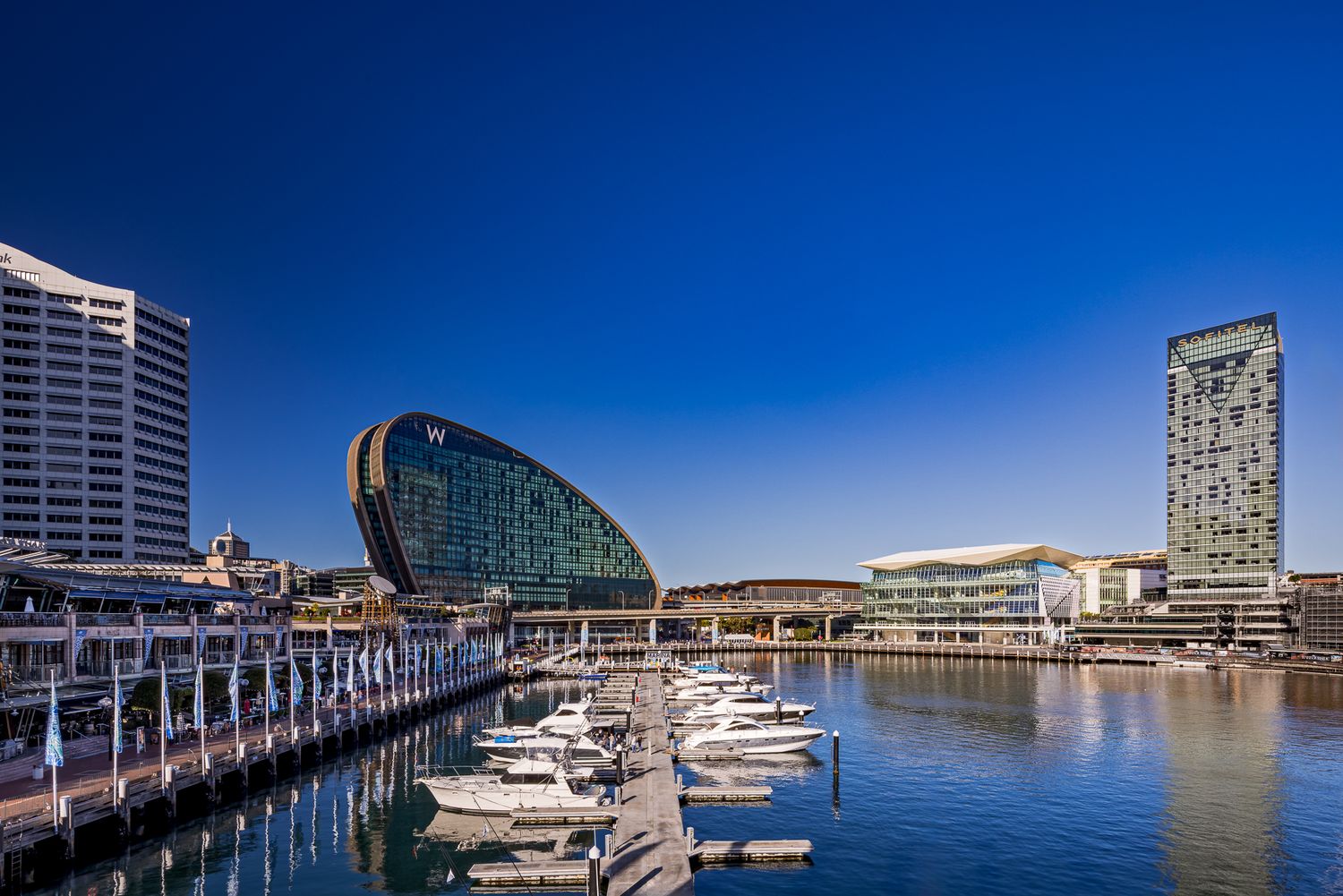 Waterfront promenade with boats docked in a marina, curved glass building and city towers reflected in calm harbour water.
