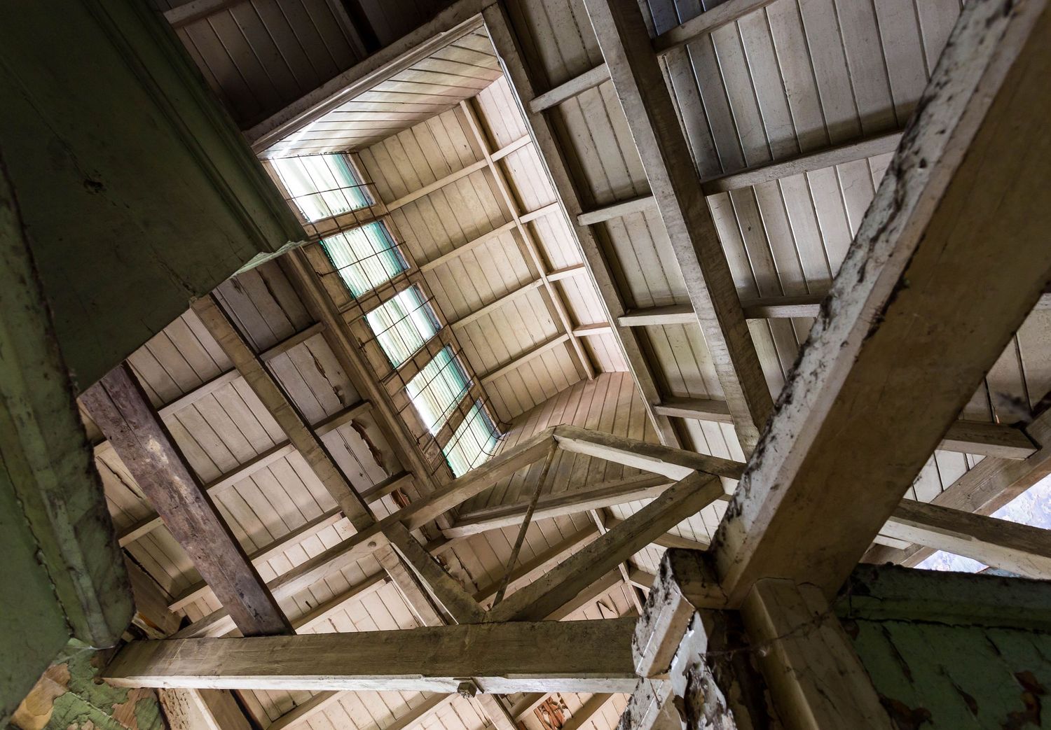 Upward view of an old timber-framed roof structure with exposed wooden beams, rafters, planked ceiling and a skylight section.