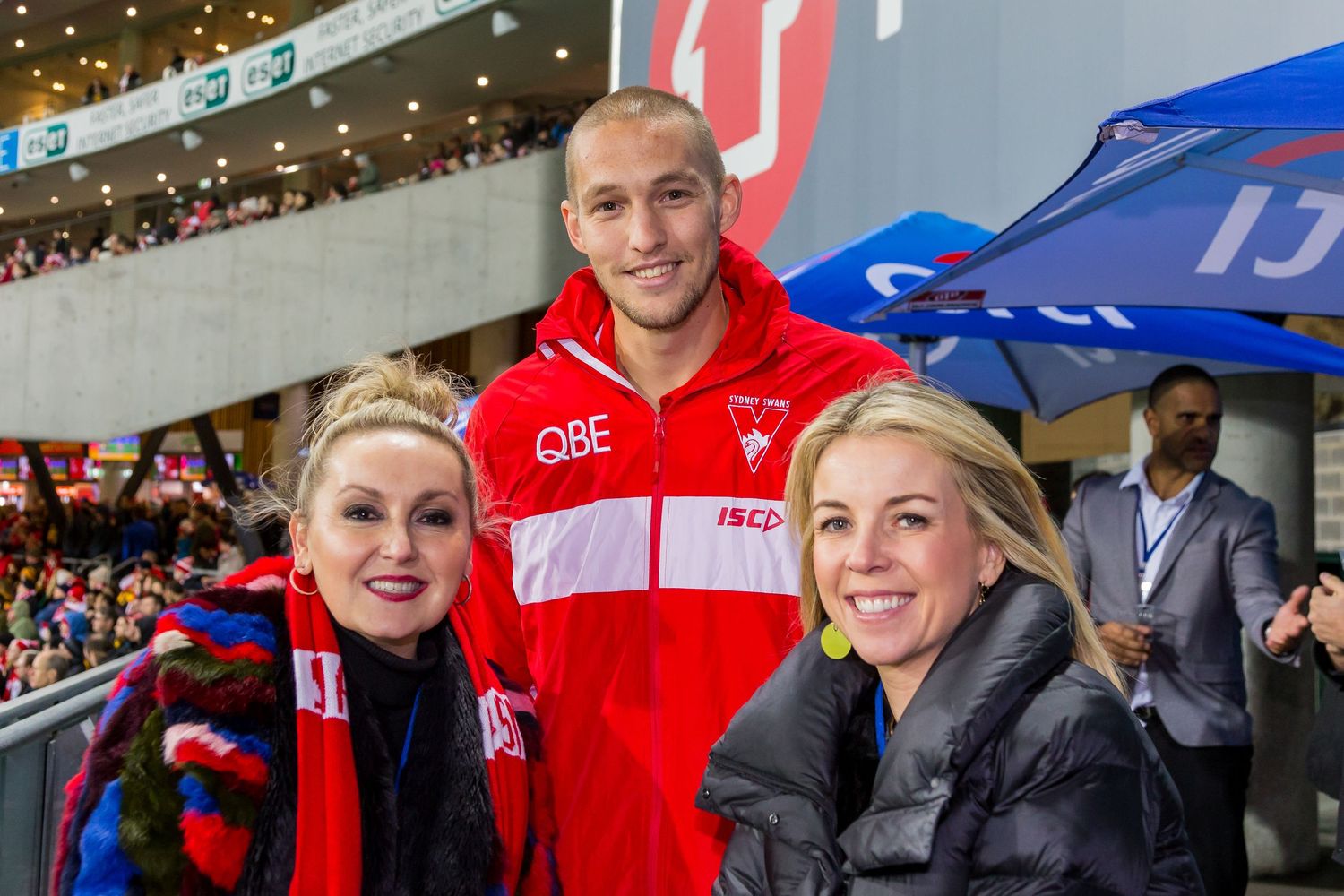 A smiling man in a red Sydney Swans jacket poses with two women at a sports stadium event.