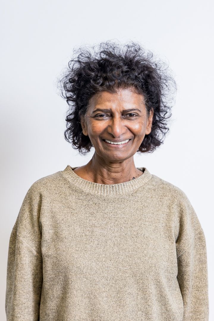A smiling woman with short curly dark hair wears a beige knit sweater and poses against a plain white background.
