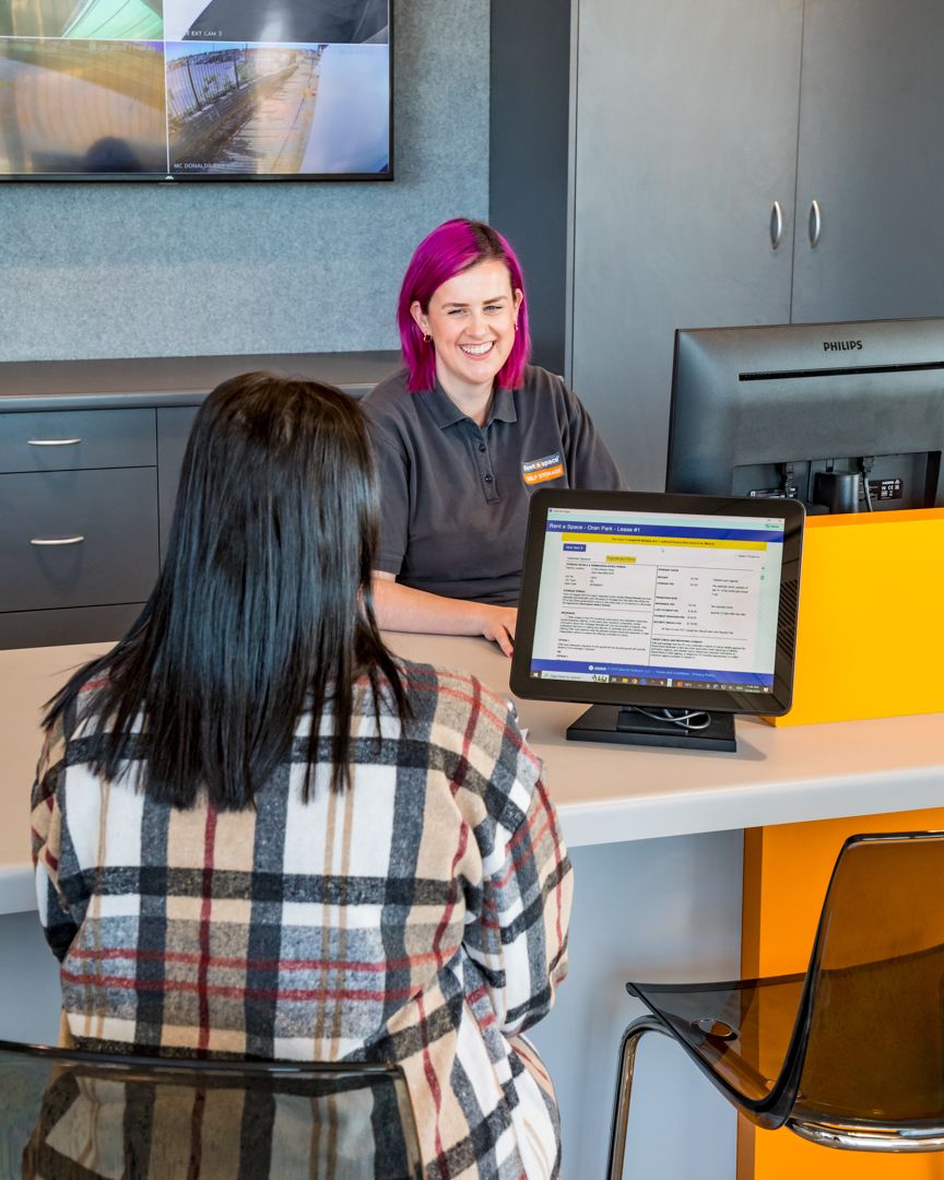 A smiling customer service employee with bright pink hair assists a woman at a counter with a tablet displaying a form.