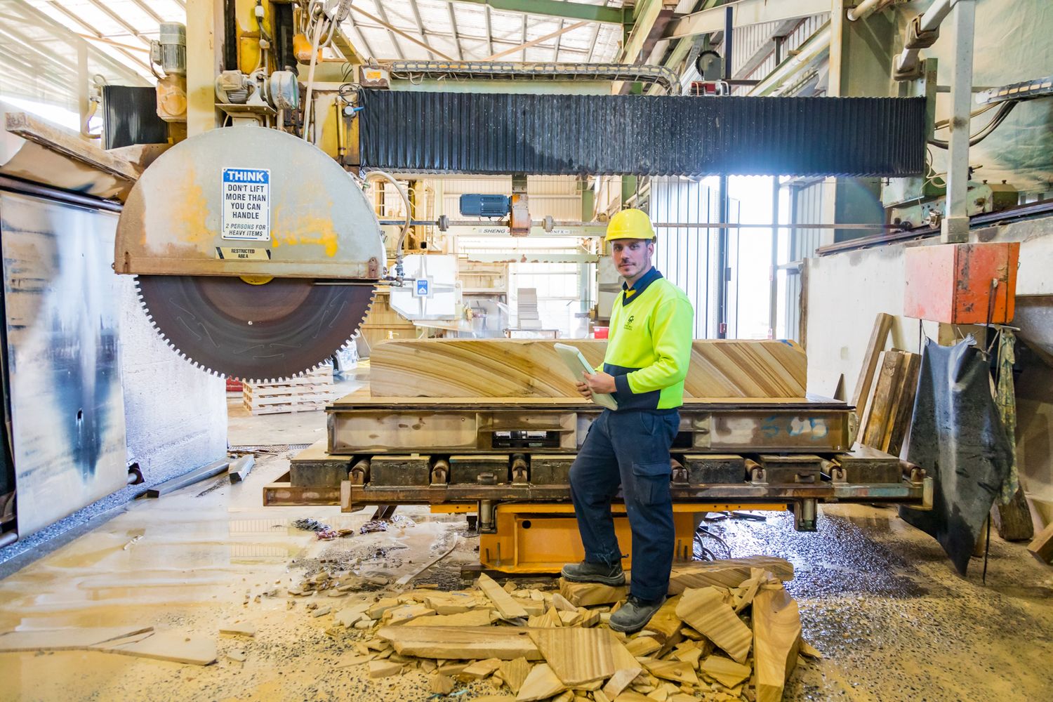 A worker in a hard hat and high-visibility jacket stands beside a large stone-cutting saw in an industrial workshop.