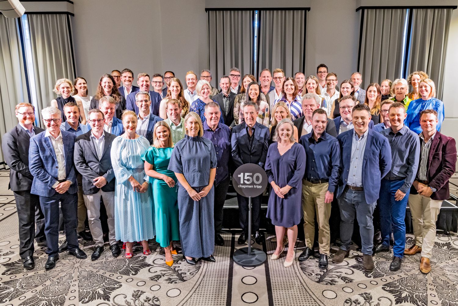 Large group of smiling professionals poses together in a conference room around a sign reading 150, marking an anniversary event.