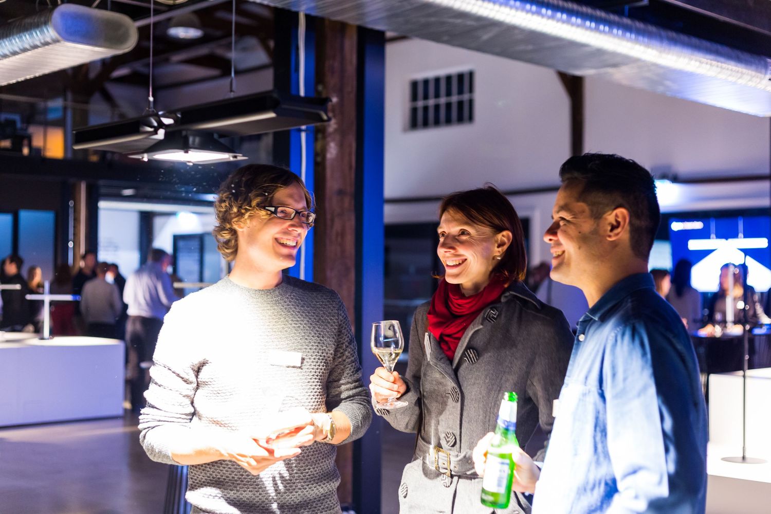 Two men and a woman chat and smile while holding drinks at an industrial-style networking event with warm lighting.