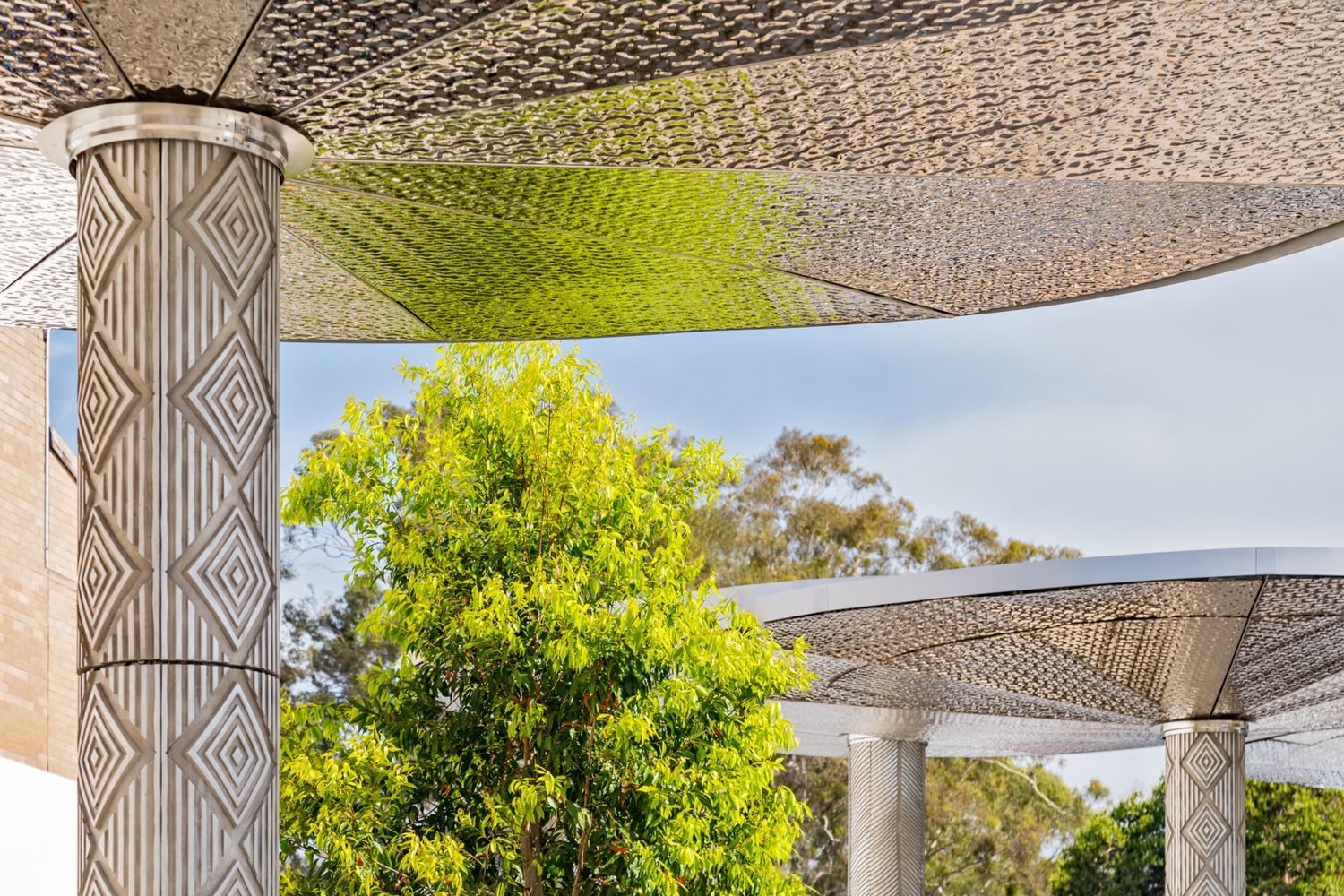 Ornate perforated metal canopies supported by patterned columns frame a green leafy tree against a soft blue sky in an outdoor shaded space.