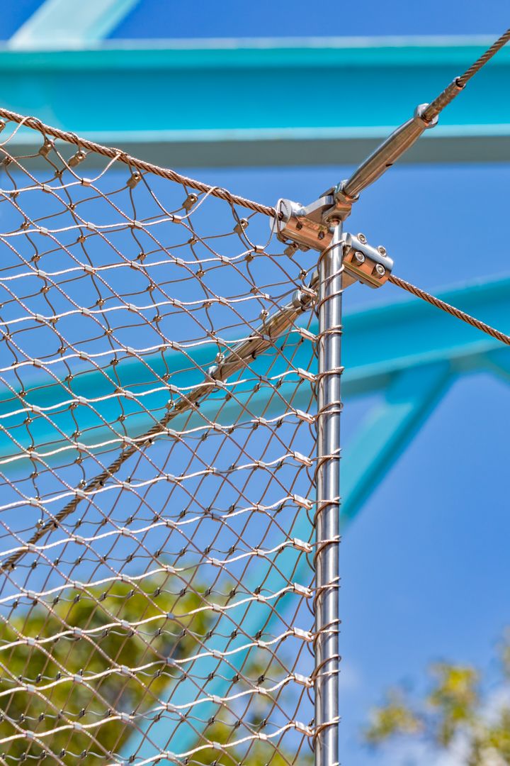 Close-up of stainless steel wire mesh netting attached to a metal post with cable fittings against a blue sky.
