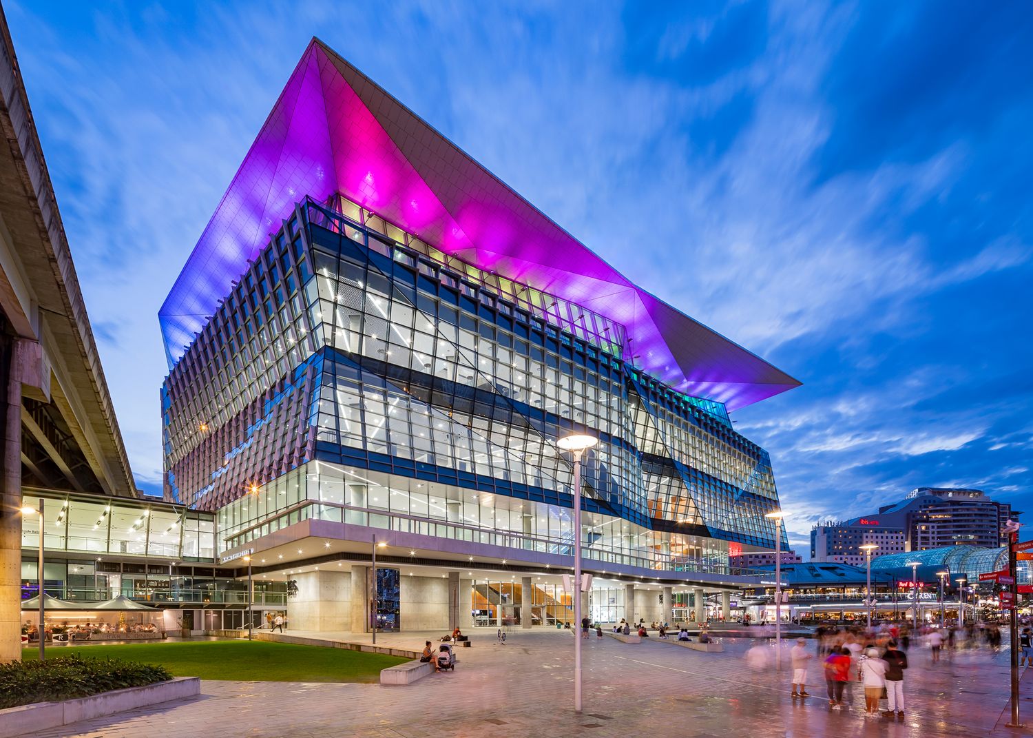 Modern glass convention centre with a glowing pink and purple angular roof lit at dusk above a landscaped public plaza.