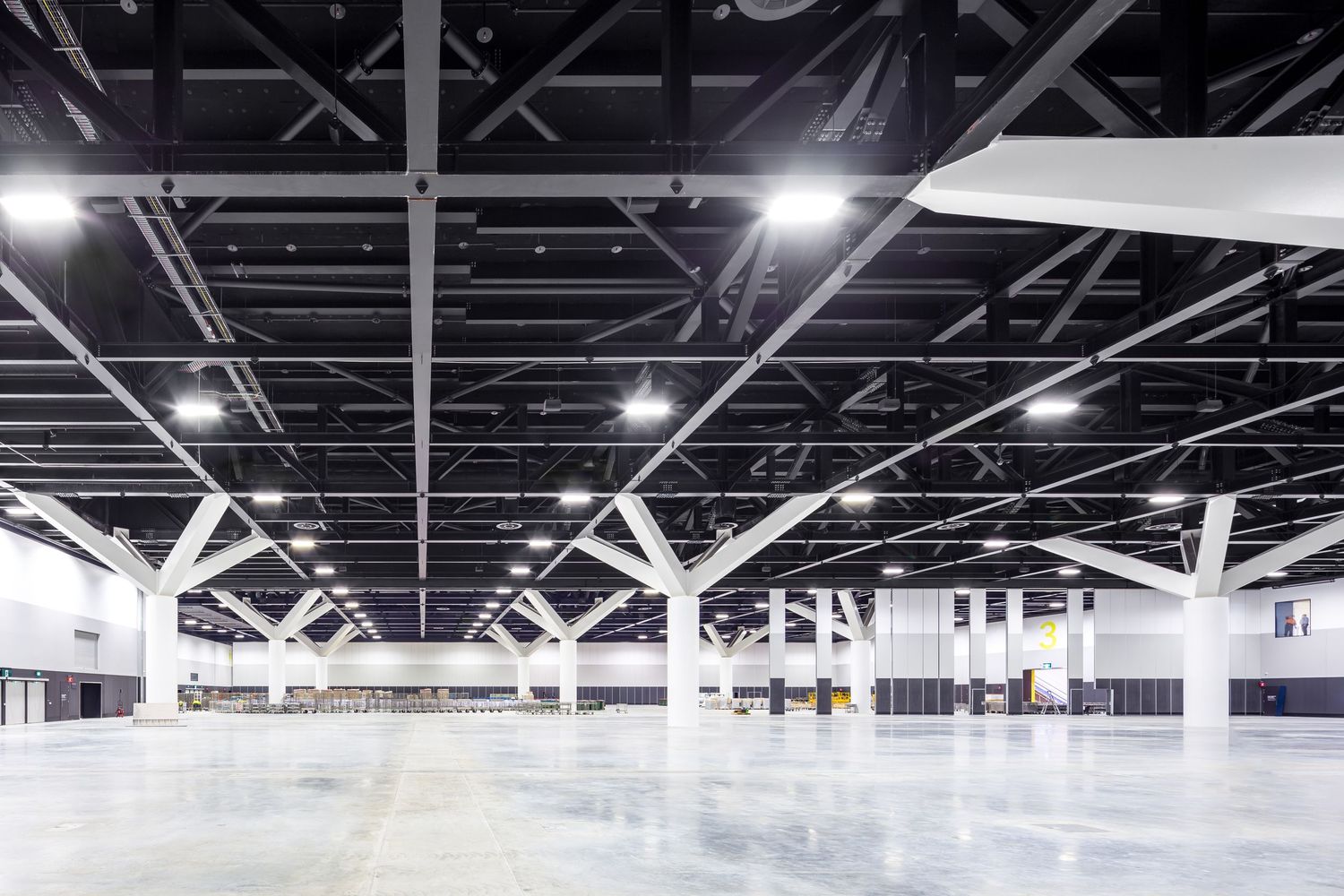 Vast empty exhibition hall with a white concrete floor, steel roof trusses and bright overhead lights ready for a large event.