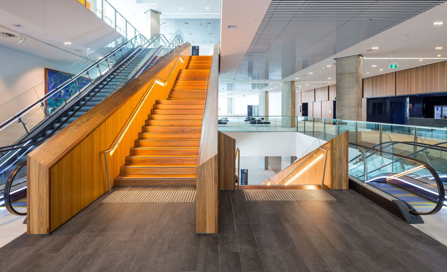 Timber-clad staircase with glowing handrails beside escalators in a bright, modern convention centre atrium.