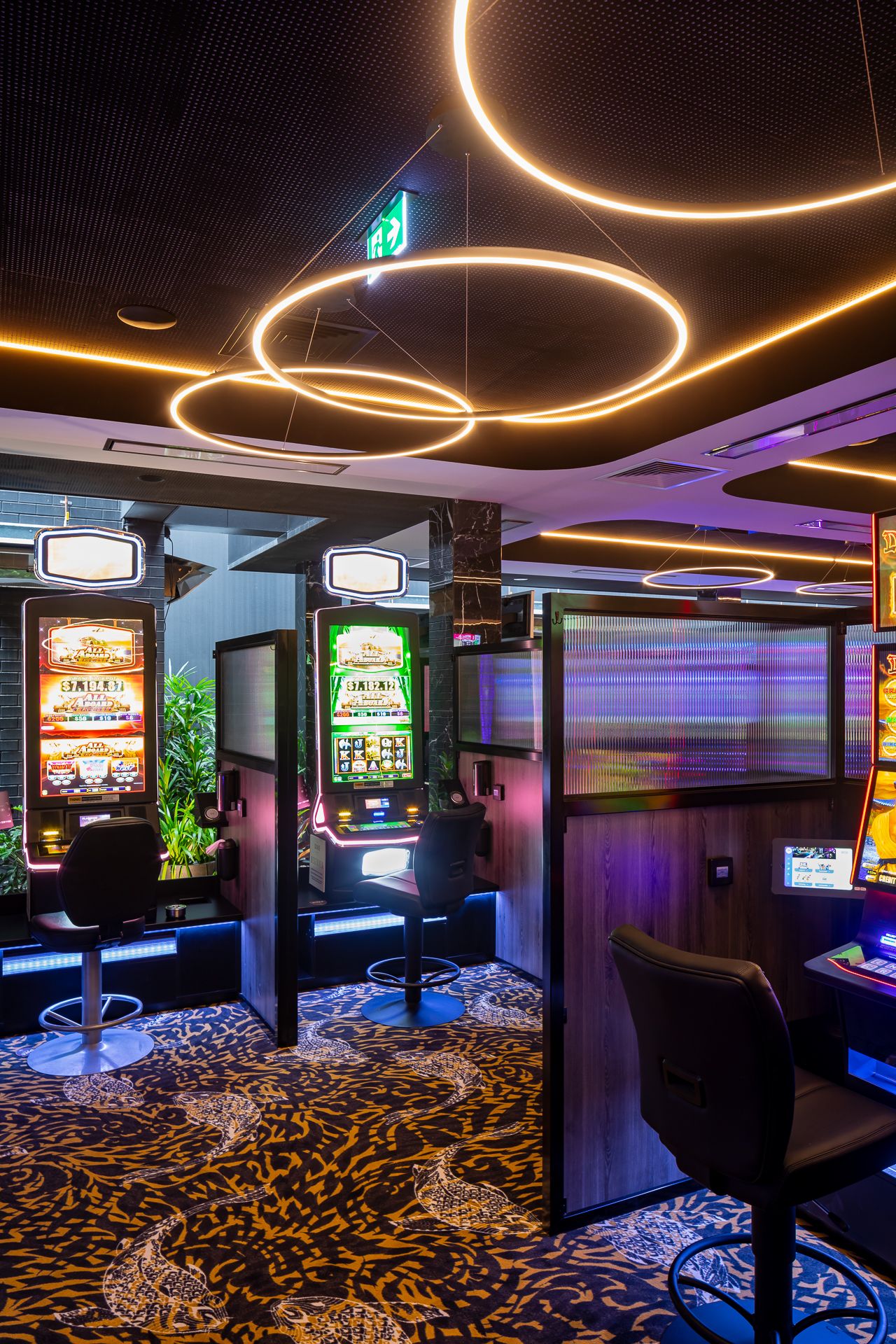 Modern casino gaming area with slot machines, black leather stools, glowing circular ceiling lights and patterned carpet.