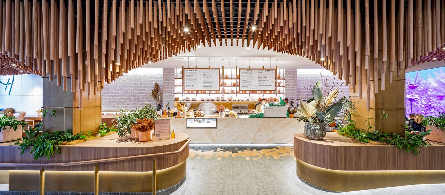 Symmetrical cafe entrance framed by arched hanging wood battens, leafy planters and a bright food display counter in the background.