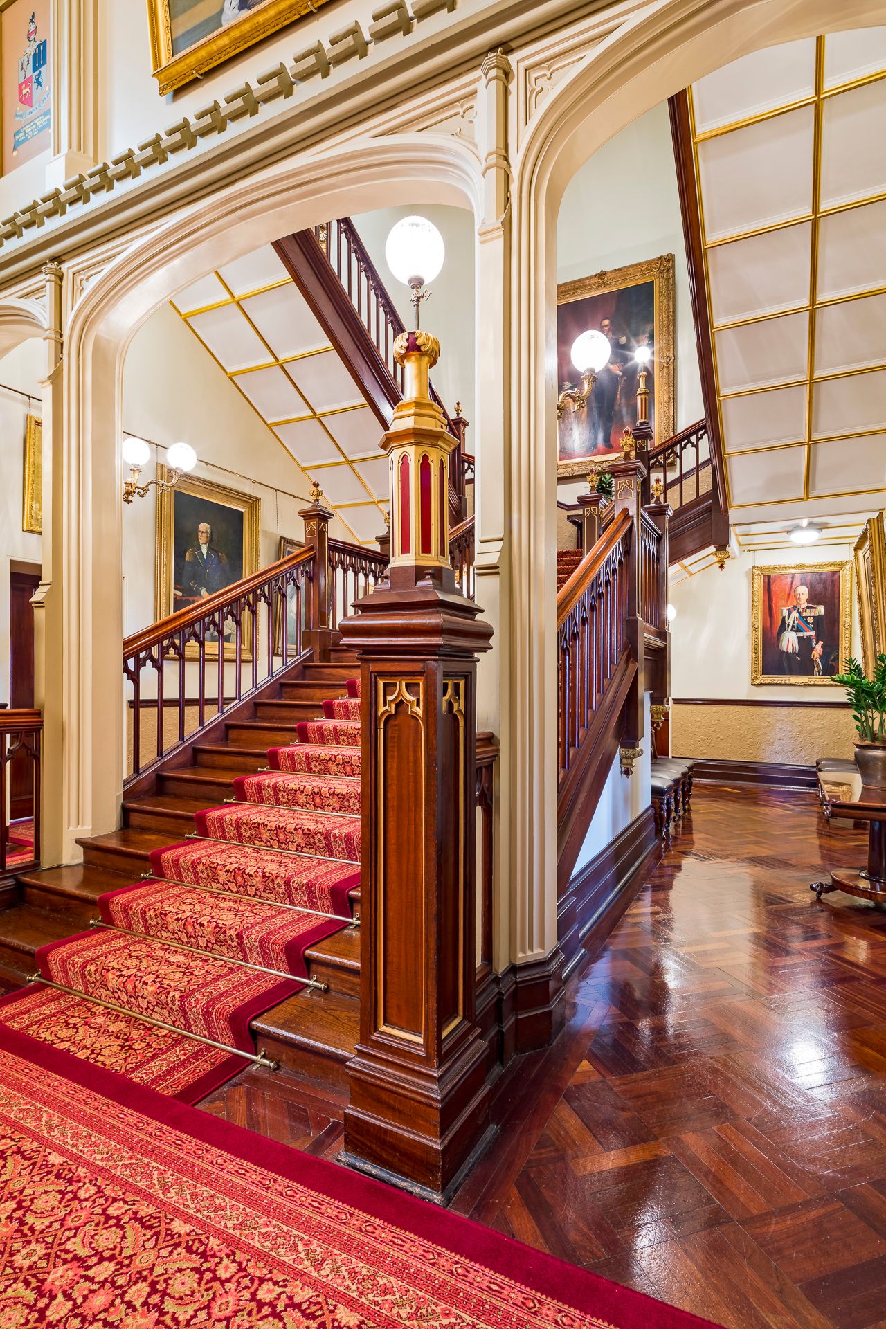 Grand heritage staircase with carved timber newel posts, red patterned carpet runner, polished parquet floor and framed portraits.