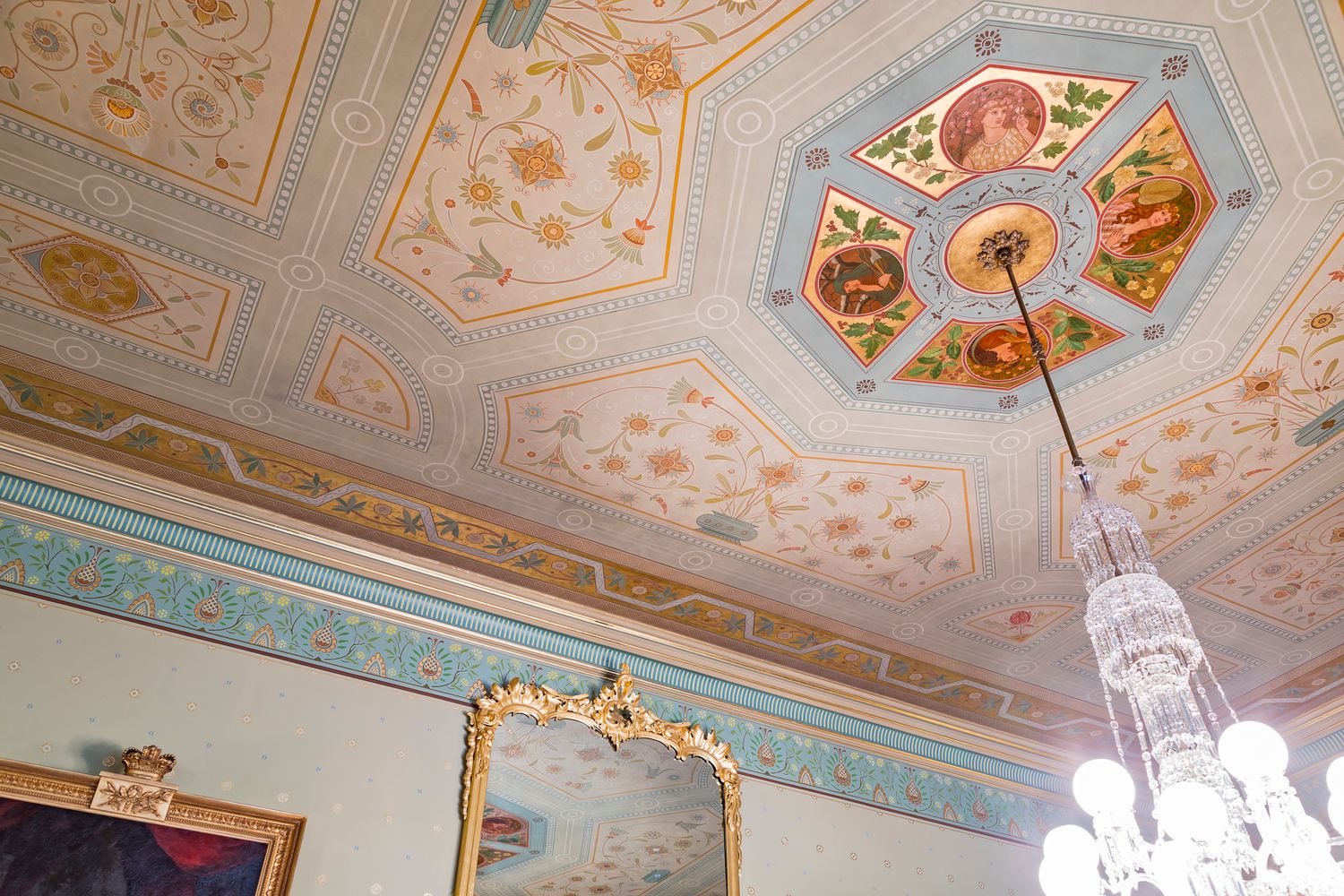 Ornate heritage ceiling with octagonal painted medallions and floral plasterwork above a crystal chandelier and gilded mirror.