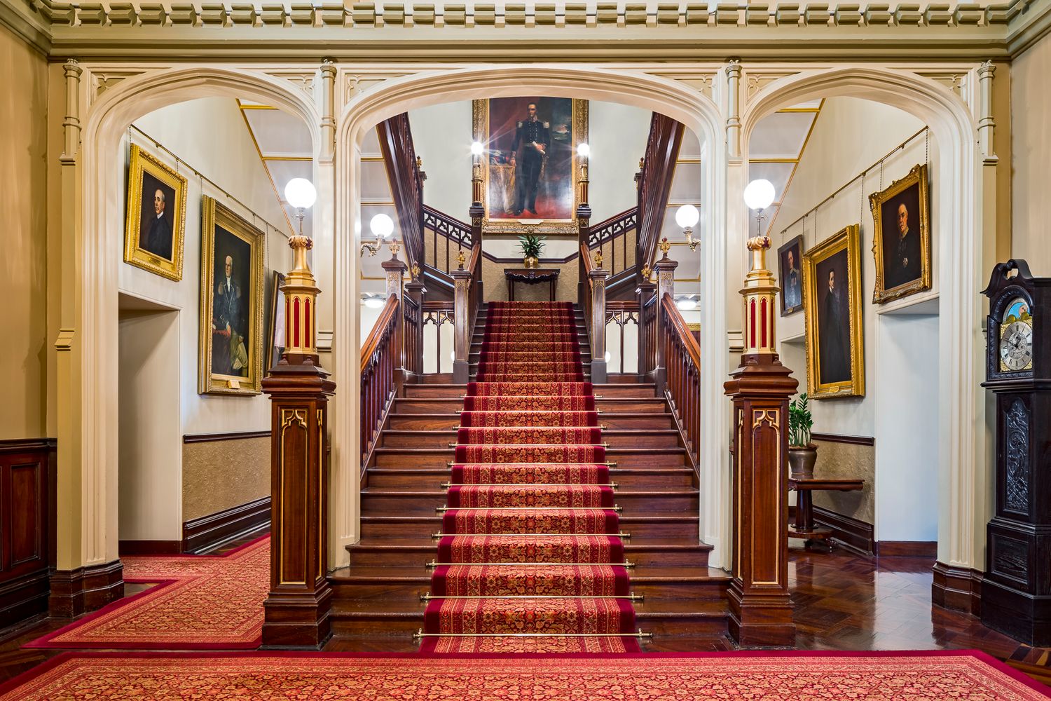 Grand historic staircase with red runner carpet, timber balustrade and framed portraits lining the walls of a heritage building.