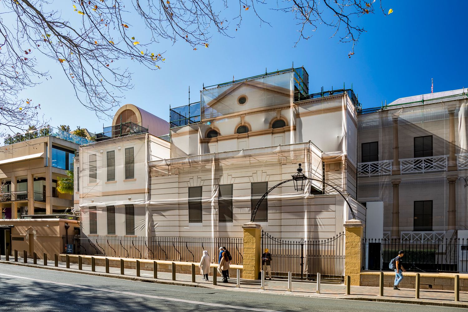 Neoclassical building facade under renovation with scaffolding, an arched lamp gate, and pedestrians on the sidewalk.