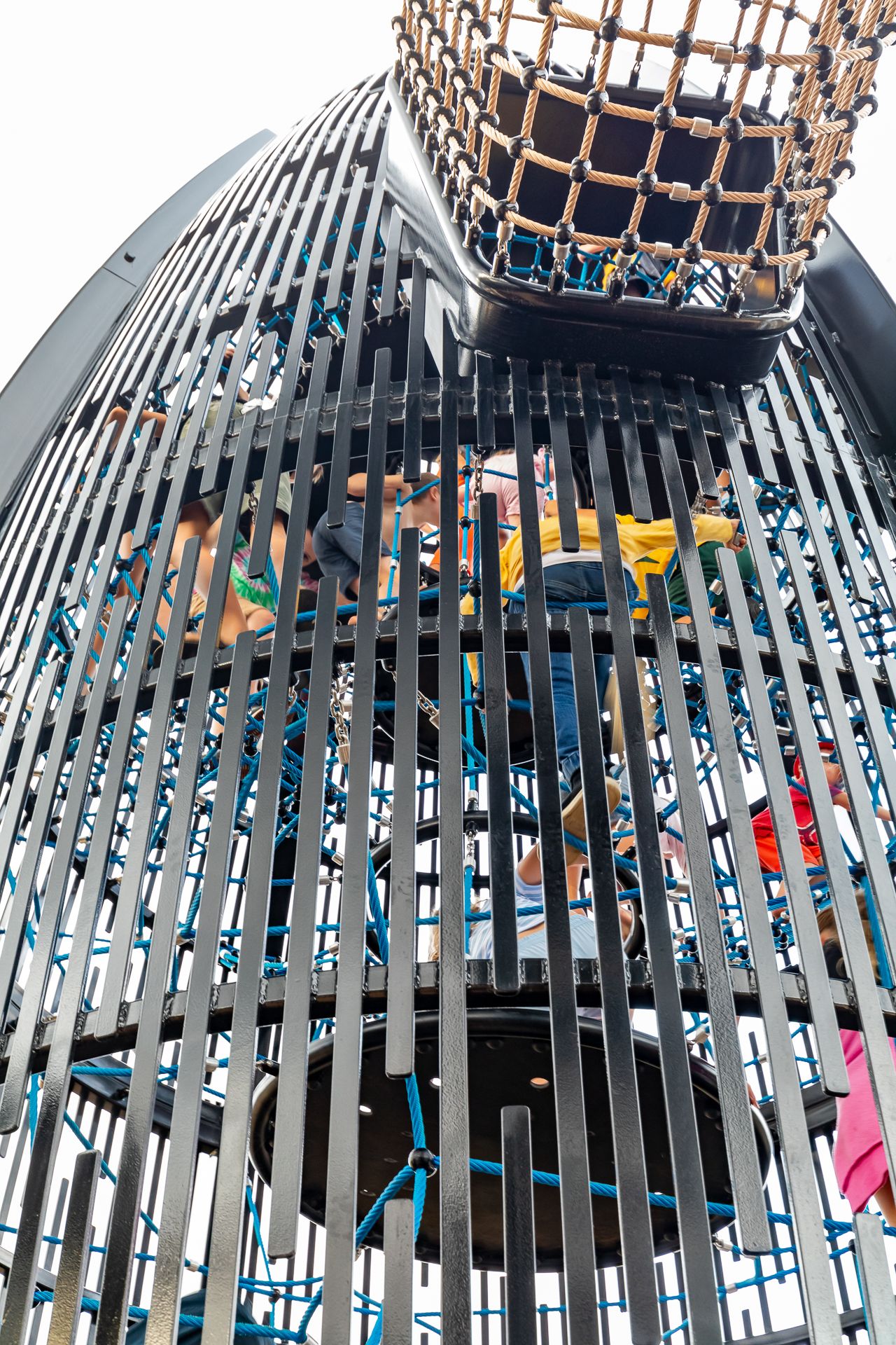 Looking up inside a tall mesh climbing tower where children scramble on rope nets beneath a woven net tunnel.
