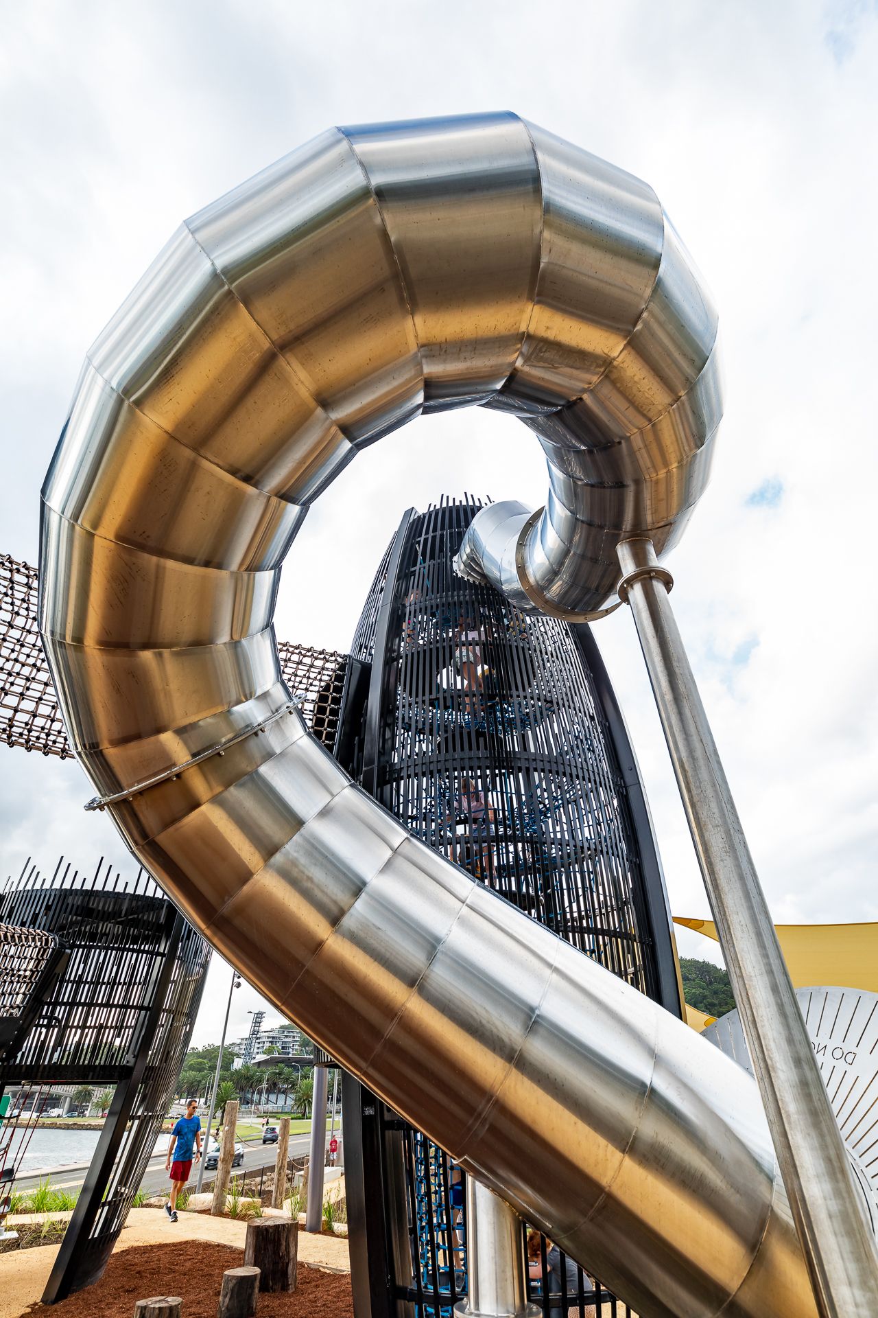 Large looping silver slide beside a lattice play tower in a waterfront park, with visitors and trees in the background.