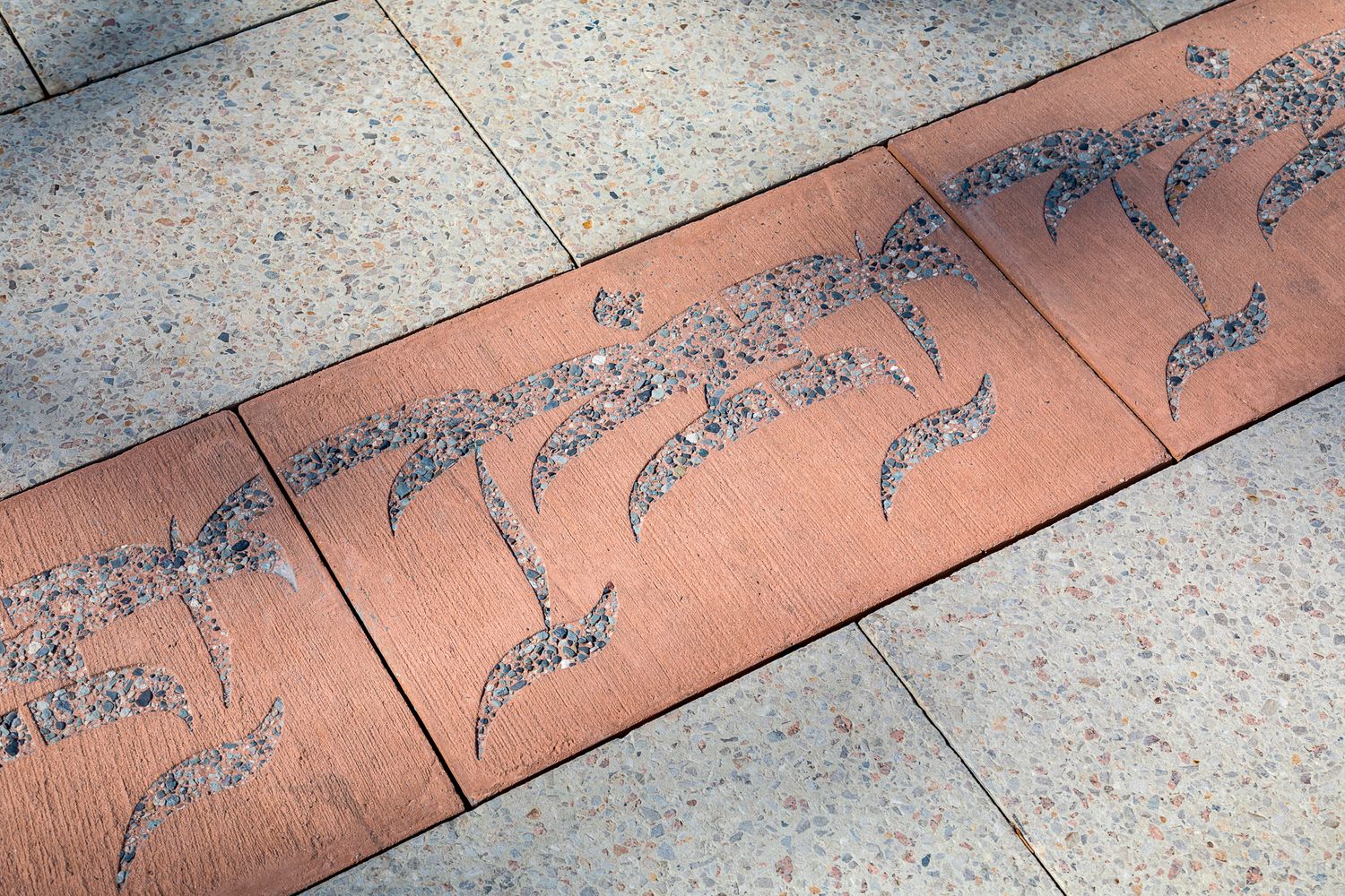 Close-up of terracotta paving pavers inlaid with textured bird-like patterns set into light speckled concrete.