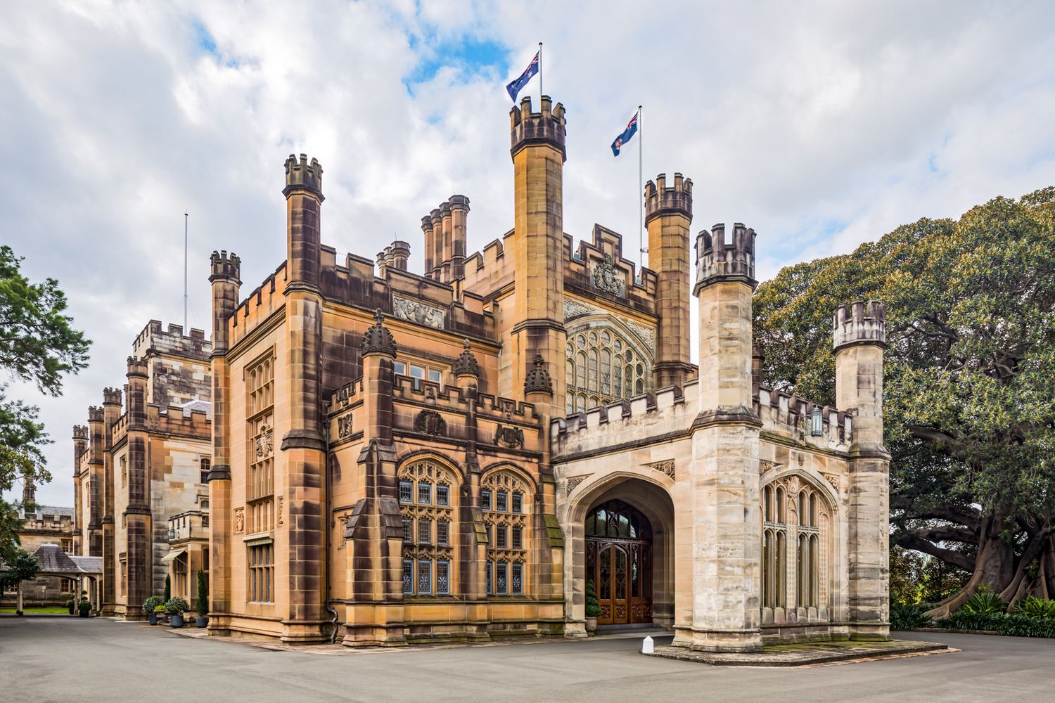 Gothic Revival sandstone castle-style building with crenellated towers, pointed arched windows and flags under a partly cloudy sky.