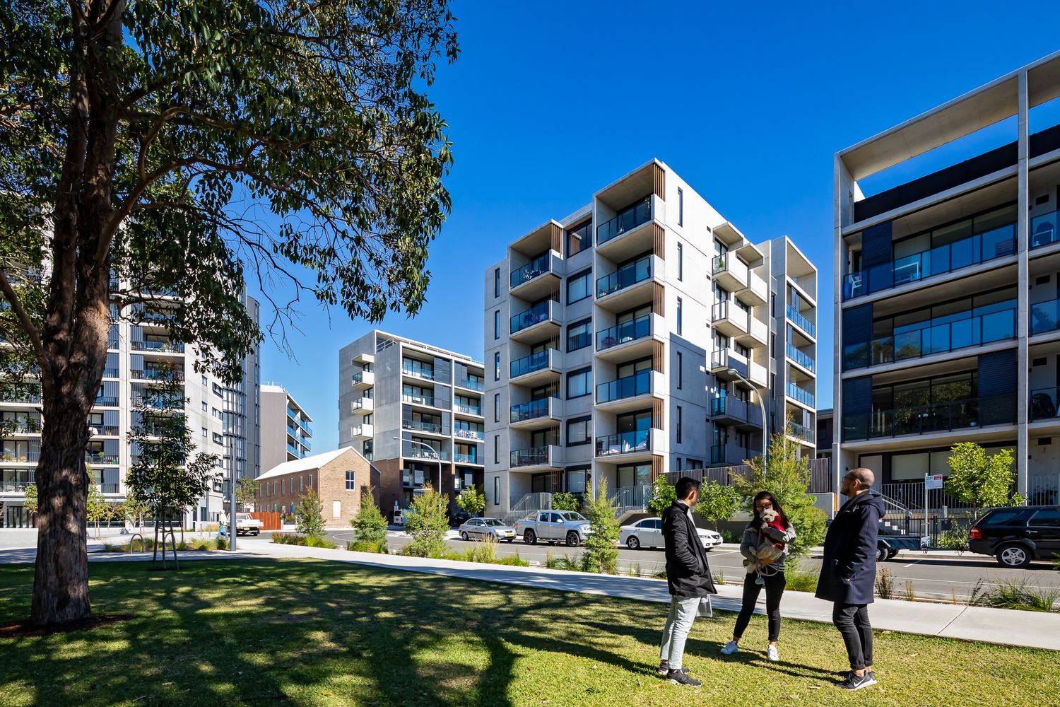 Three people stand on a sunny green lawn in front of modern multi-story apartment buildings with balconies, shaded by large trees.