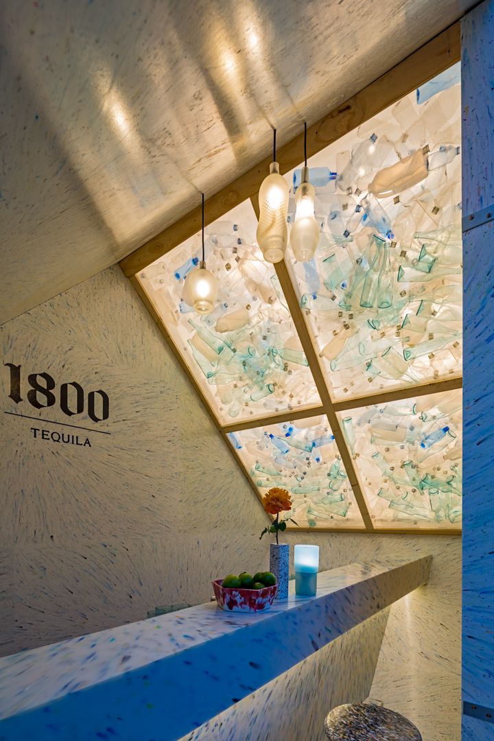 Interior of a 1800 Tequila bar with a slanted skylight made of recycled glass bottles, pendant lights and a white counter with a flower.