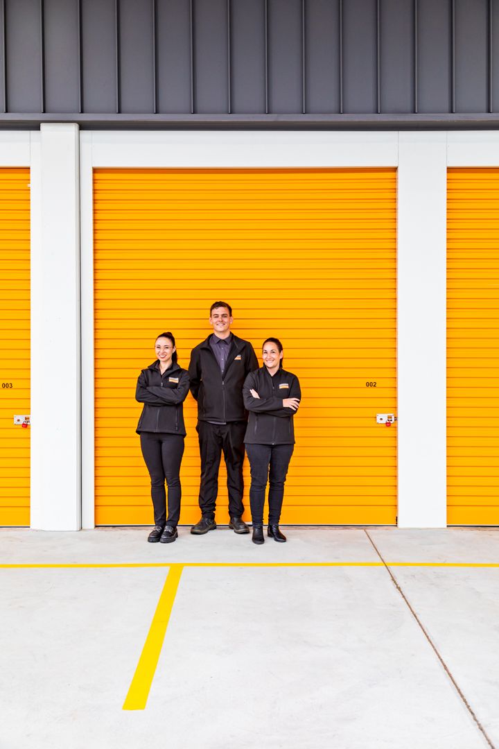 Three staff members in black uniforms standing with arms crossed in front of bright orange self-storage unit doors.