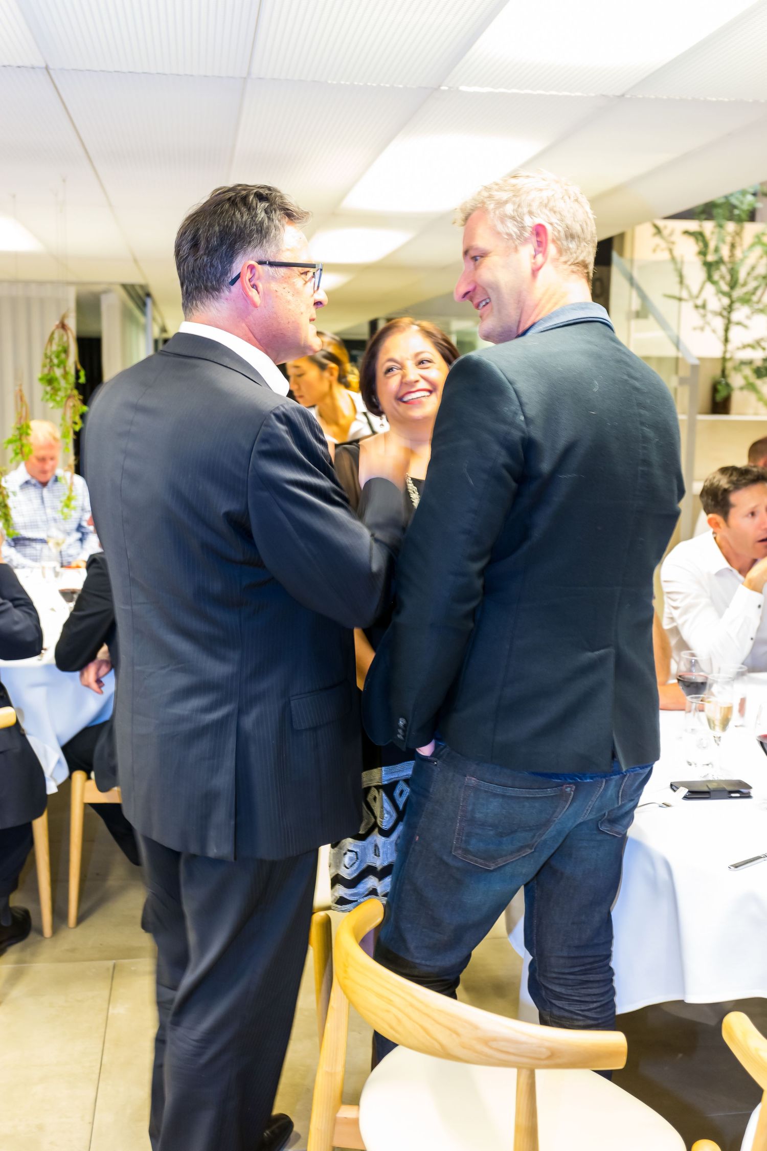 Two men in dark jackets greet each other warmly beside a smiling woman at a busy indoor dining event with guests seated at tables.