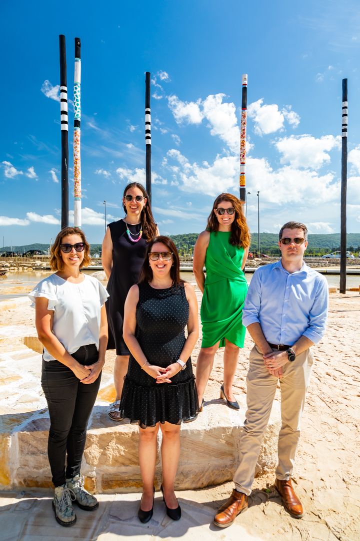 Four women and a man pose on a sandstone ledge by the water, with tall decorated poles and blue sky behind them.