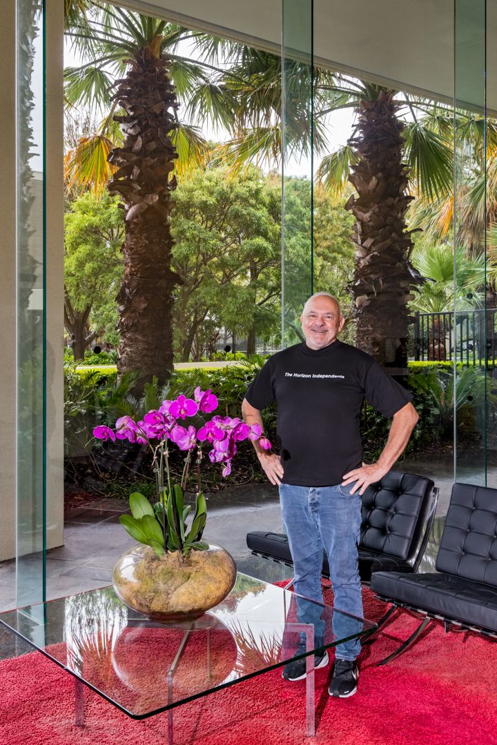 A smiling man in a black t-shirt and jeans stands beside a glass table with a purple orchid, with palm trees visible through large windows.
