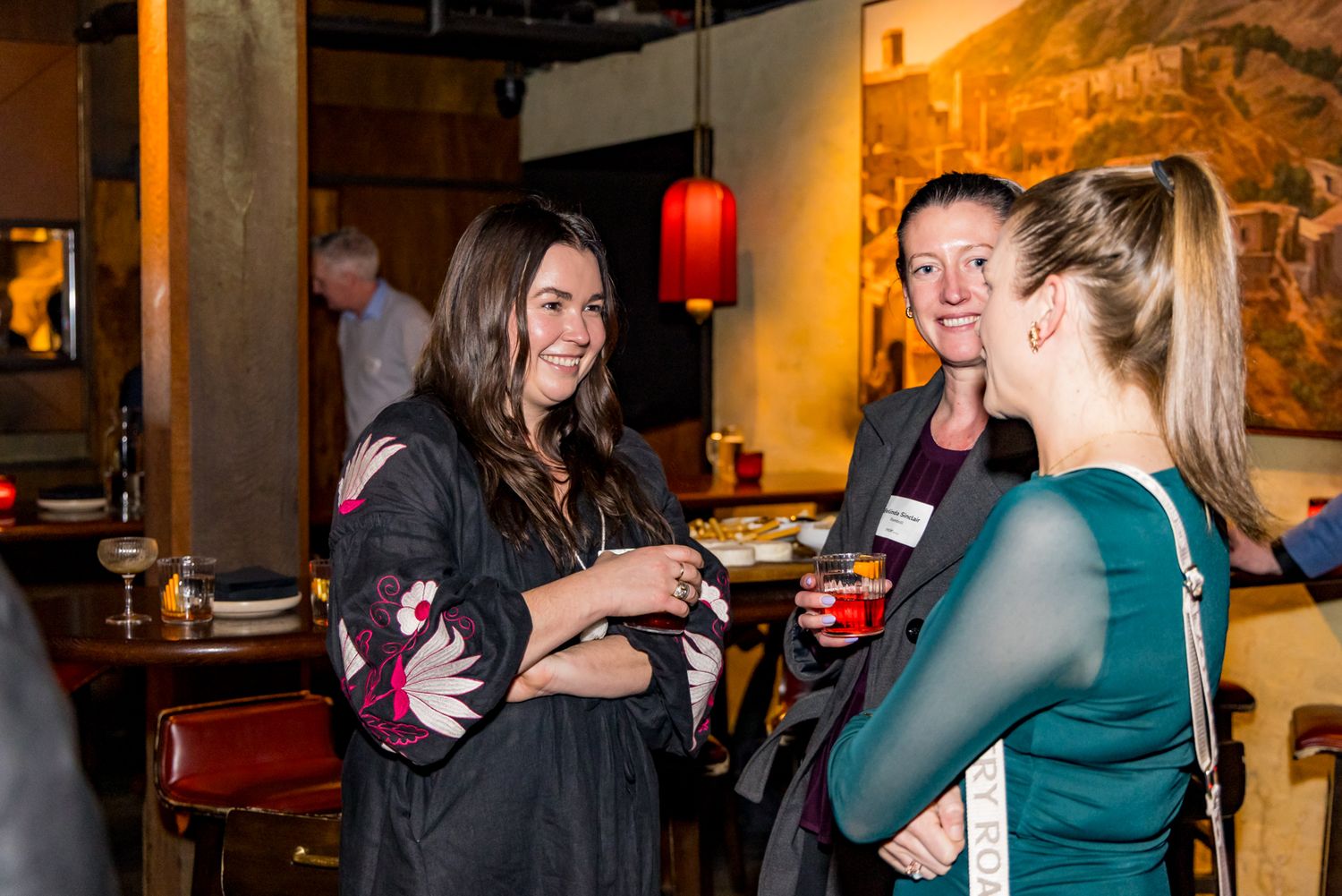 Three women chat and smile with drinks in hand at a warmly lit restaurant or bar during a casual networking event.