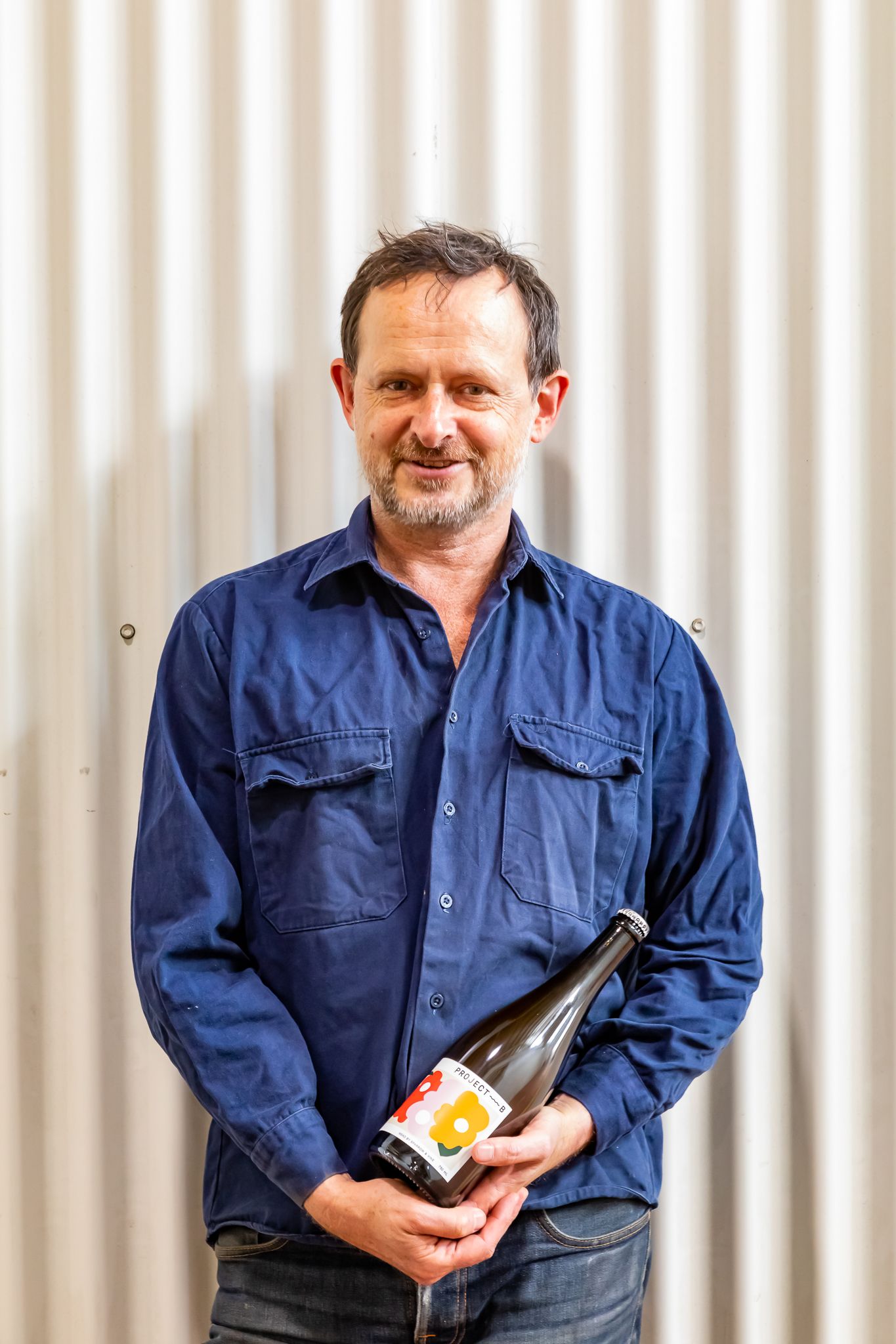 A smiling man with stubble wears a blue button-up shirt and holds a bottle of wine against a corrugated metal wall.