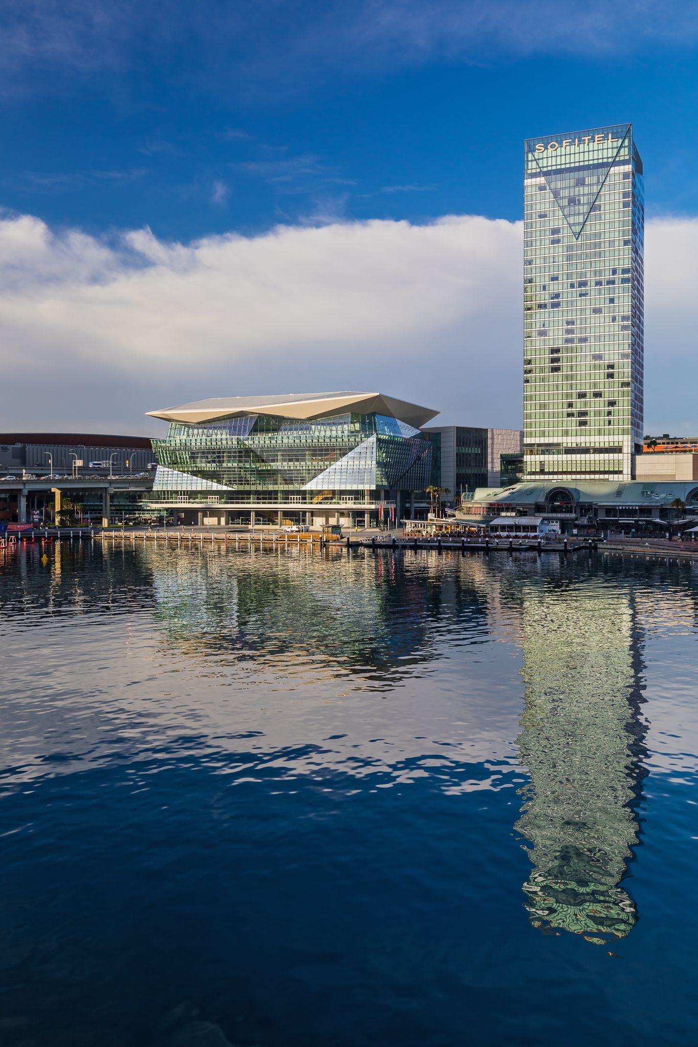 Glass convention centre and tall skyscraper reflected in calm harbour waters beneath a blue sky with soft clouds.