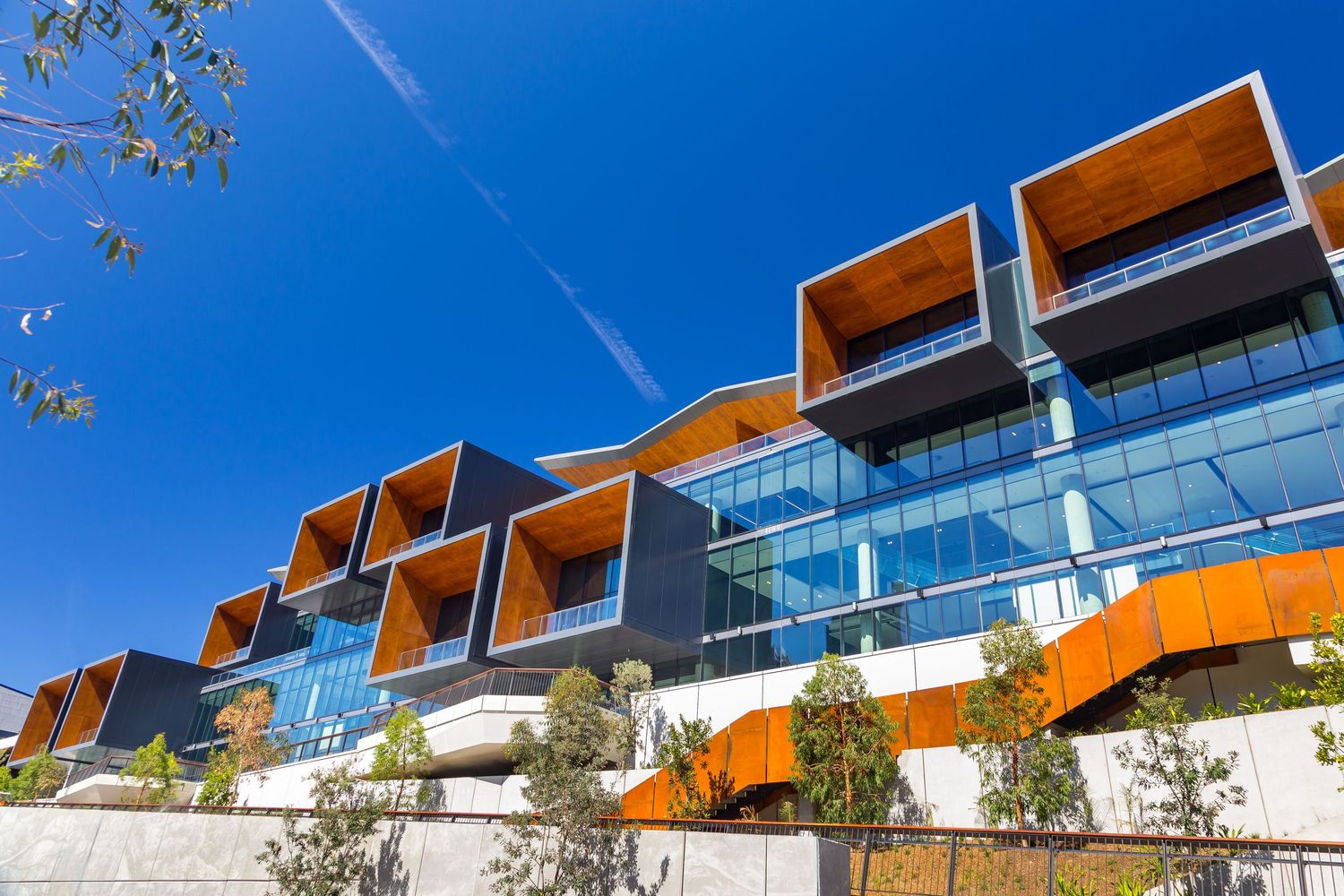 Contemporary building with angular timber-lined balconies, floor-to-ceiling glass and a curved orange walkway framed by green leaves.