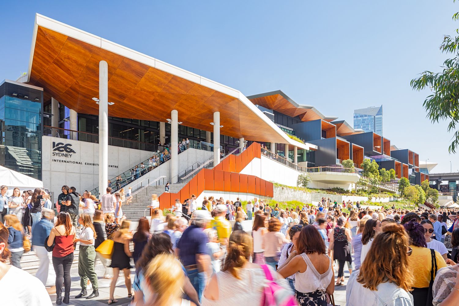 Crowds gather outside a contemporary convention centre with a sweeping timber canopy roof and orange stairs on a sunny day.