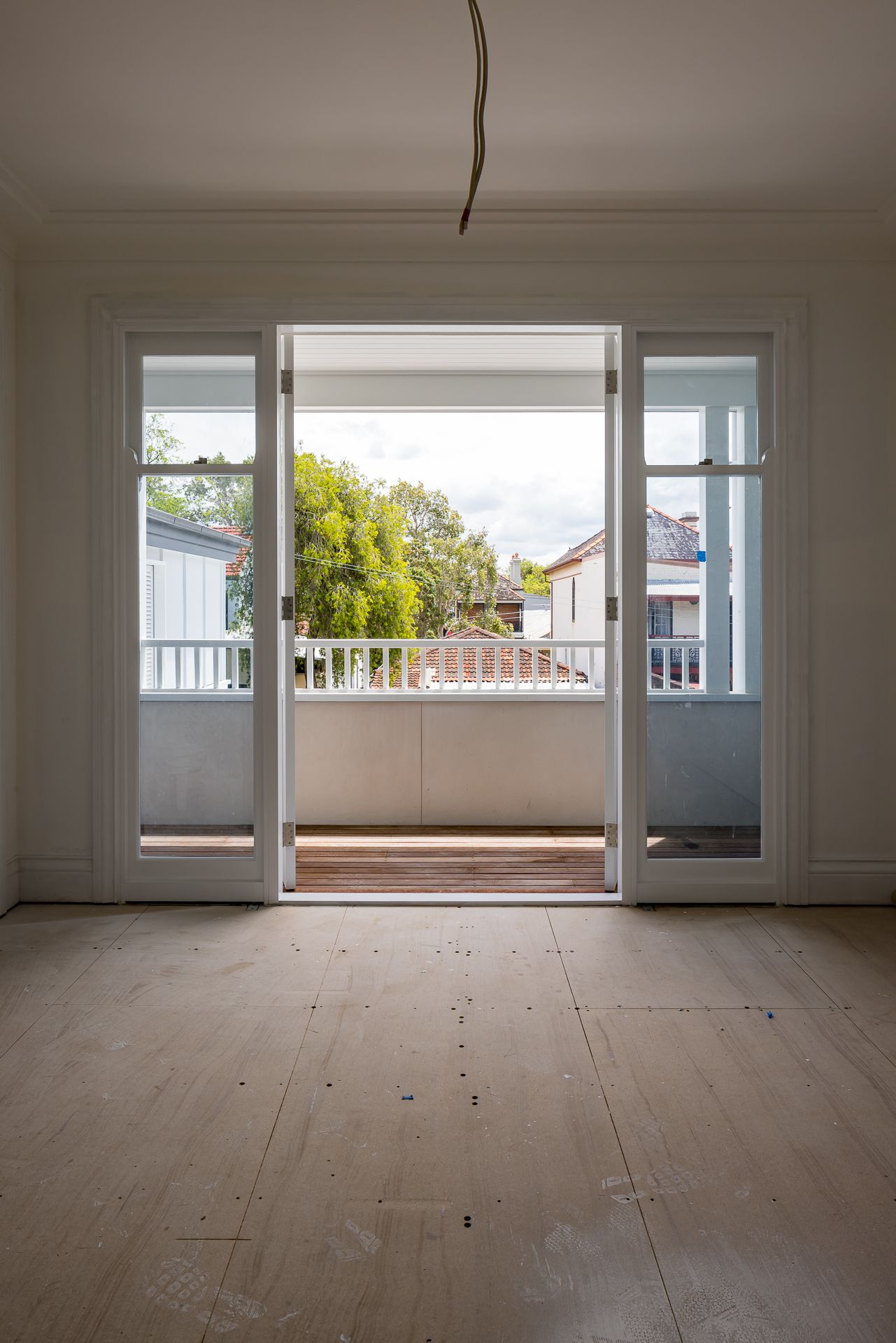 Empty room with bare plywood floors and open French doors leading to a timber balcony with white railing and leafy views.