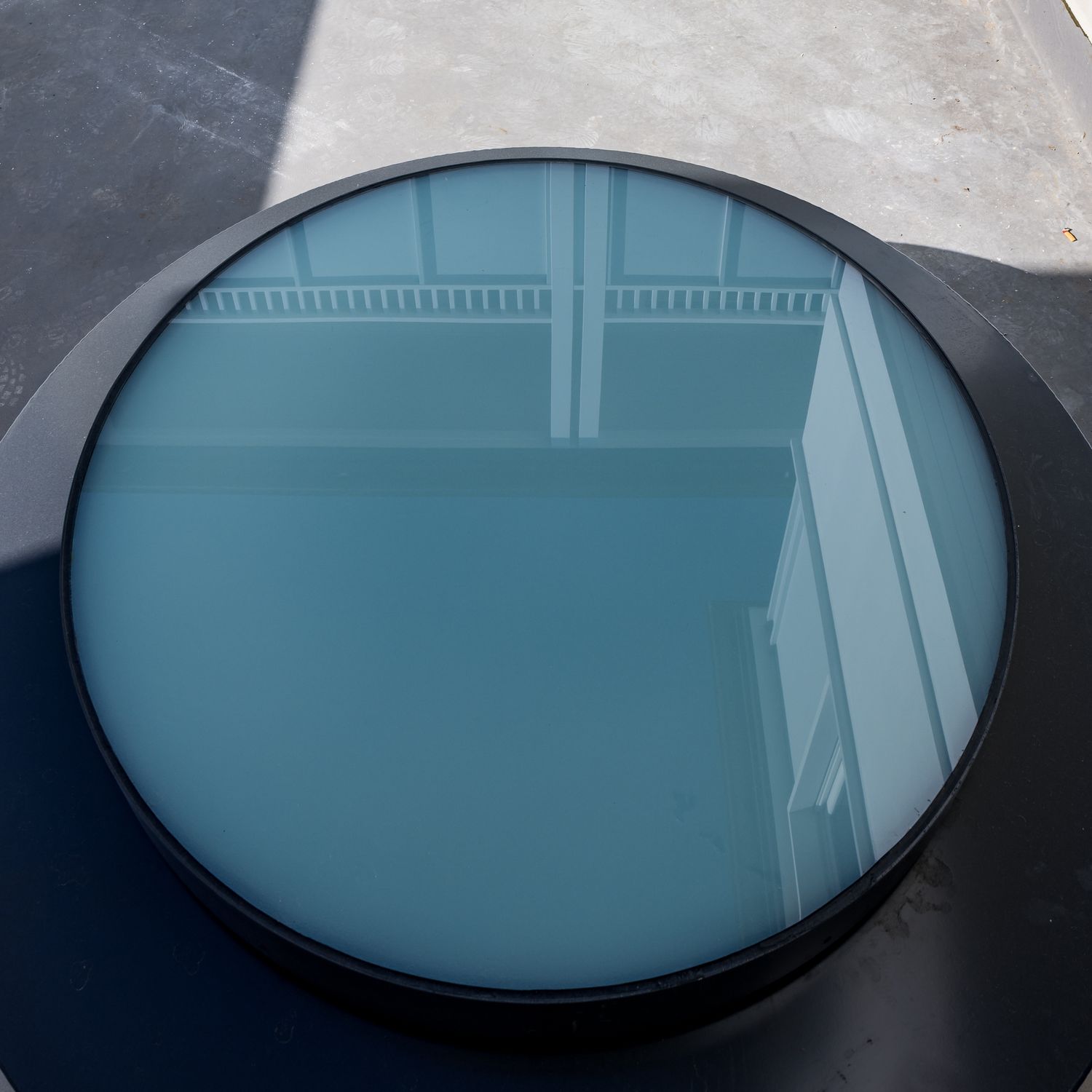 Round skylight with dark frame reflecting the blue-tinted glass and a white building facade above a concrete surface.
