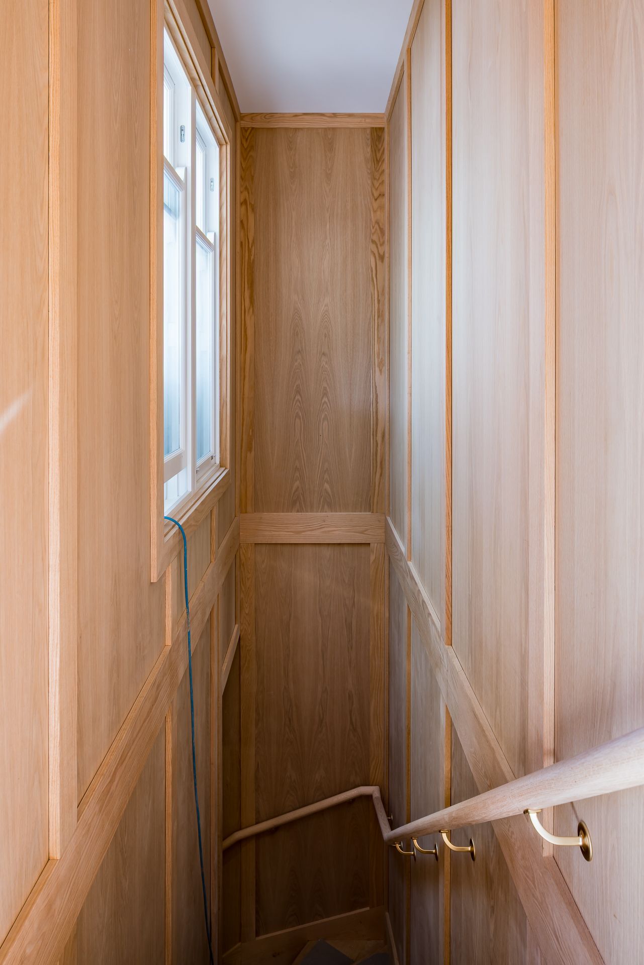 Narrow staircase lined with timber panelling, a curved wooden handrail and a small window during home renovation work.