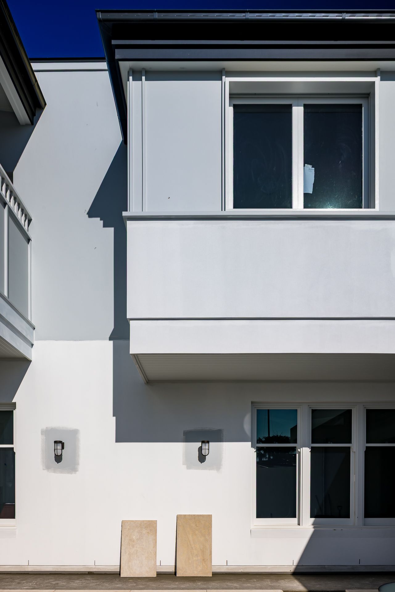 Rear exterior of a modern white two-storey townhouse with an upper balcony, black-framed windows, wall lights and stone tiles.