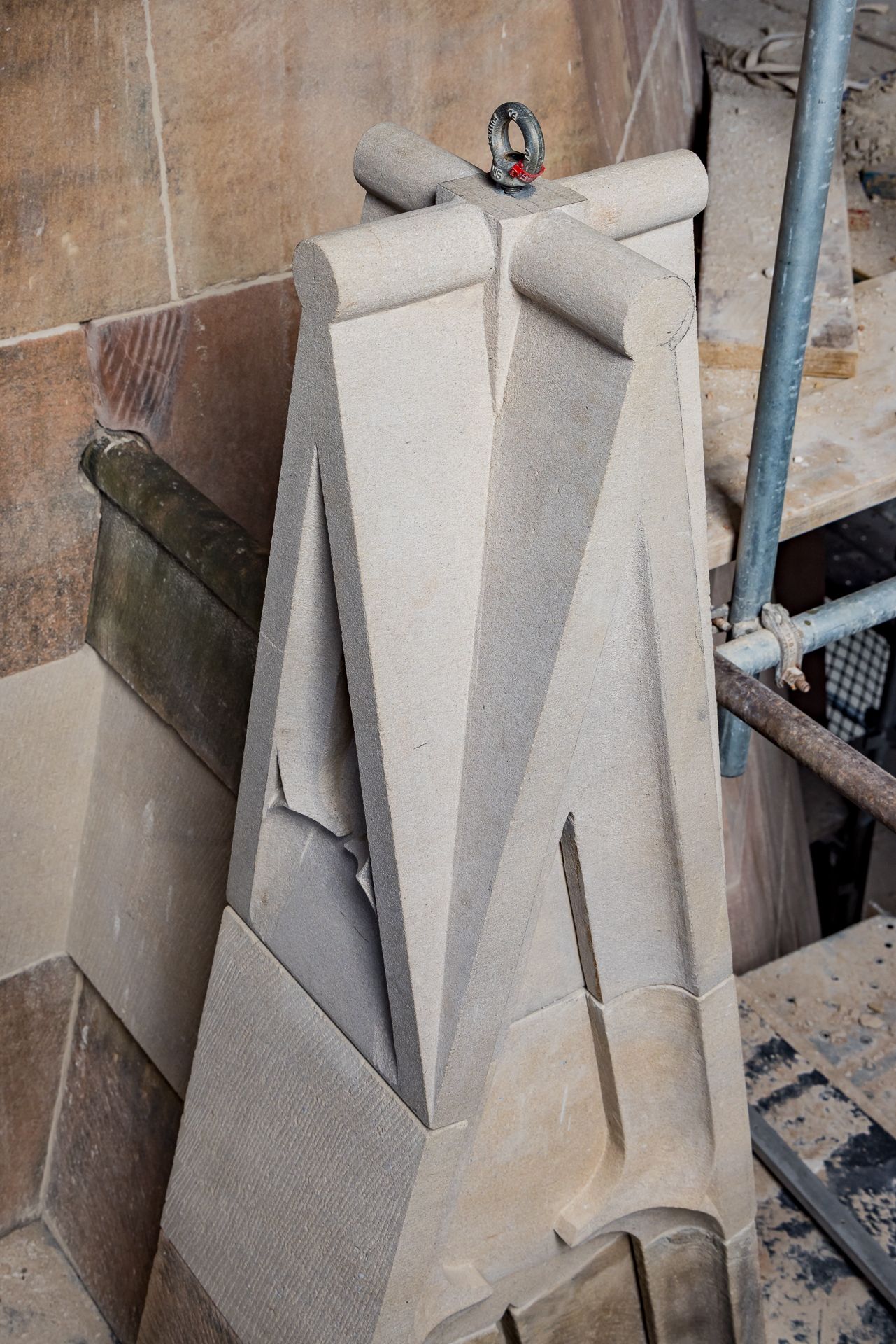 Freshly cut sandstone pinnacle fitted with a lifting eye bolt sits against old stonework beside scaffolding poles.