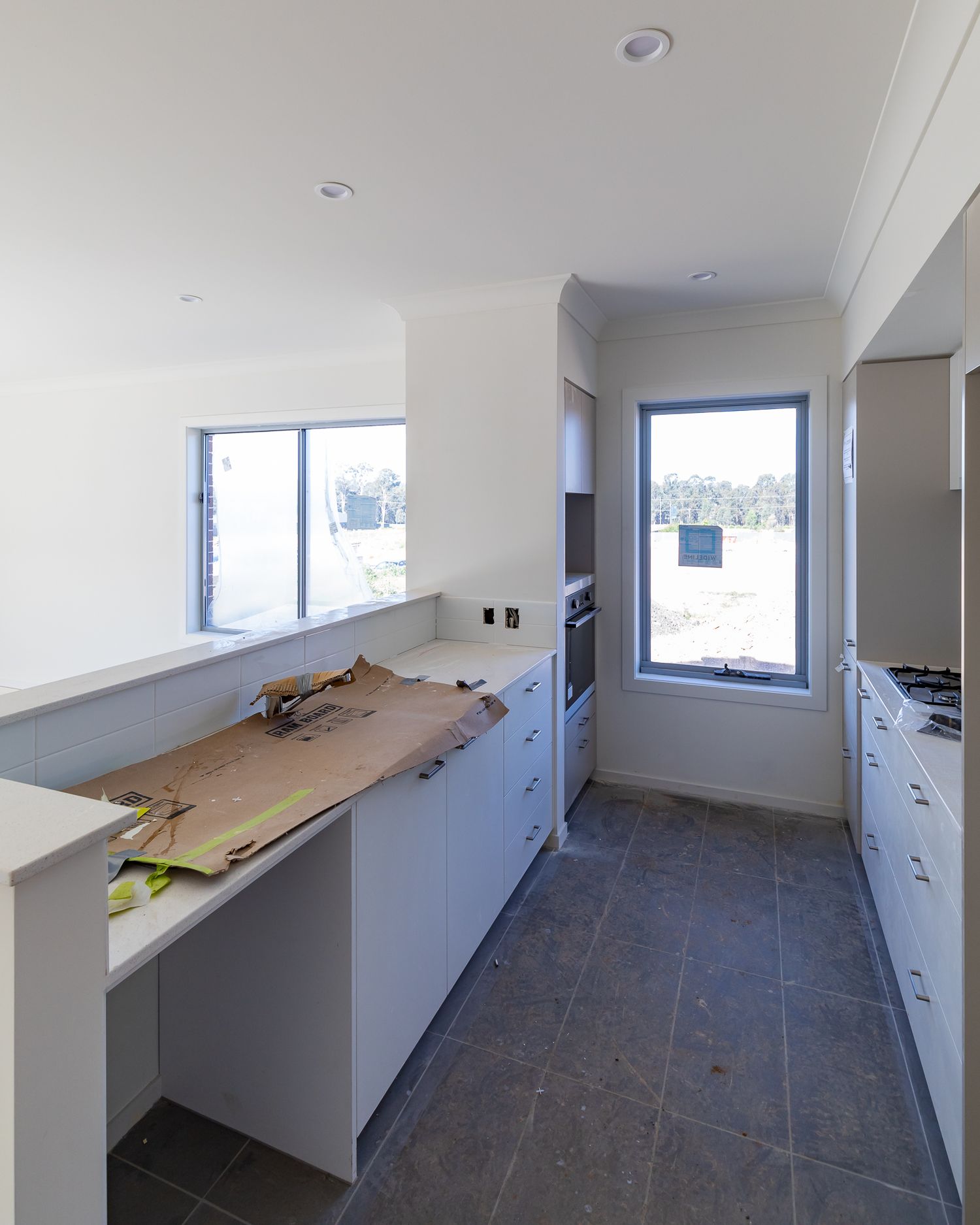 Unfinished townhouse kitchen with white cabinetry, built-in oven, protected benchtop and dark tiled floor beside large windows.