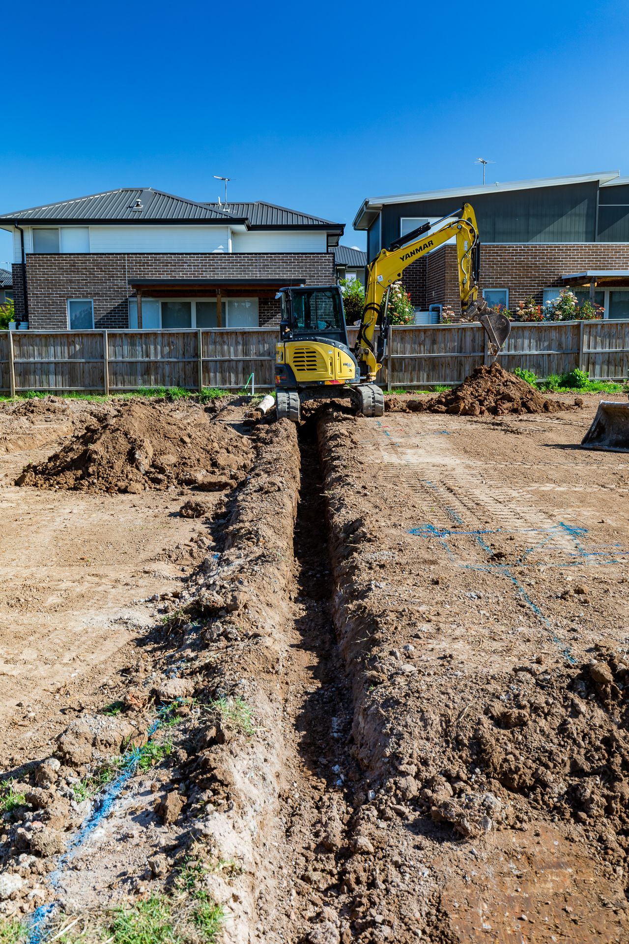 A yellow mini excavator stands behind a freshly dug trench on a residential construction site with houses in the background.