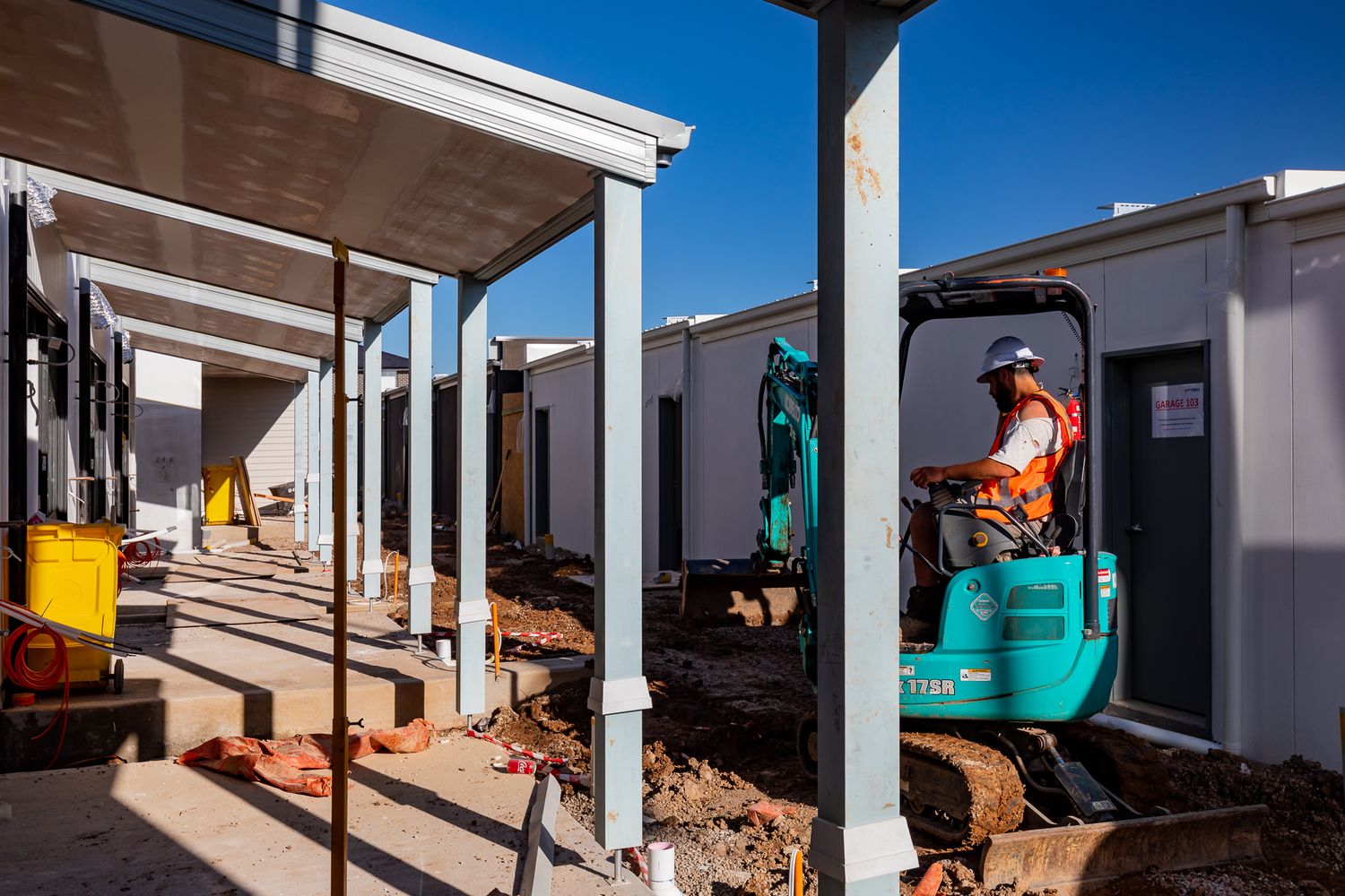 Worker in an orange vest operates a teal mini excavator beside new modular homes with covered verandahs at a building site.