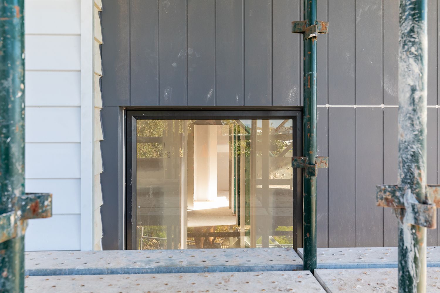 Close-up of a construction site window in dark cladding framed by scaffolding poles, with a bright interior visible.