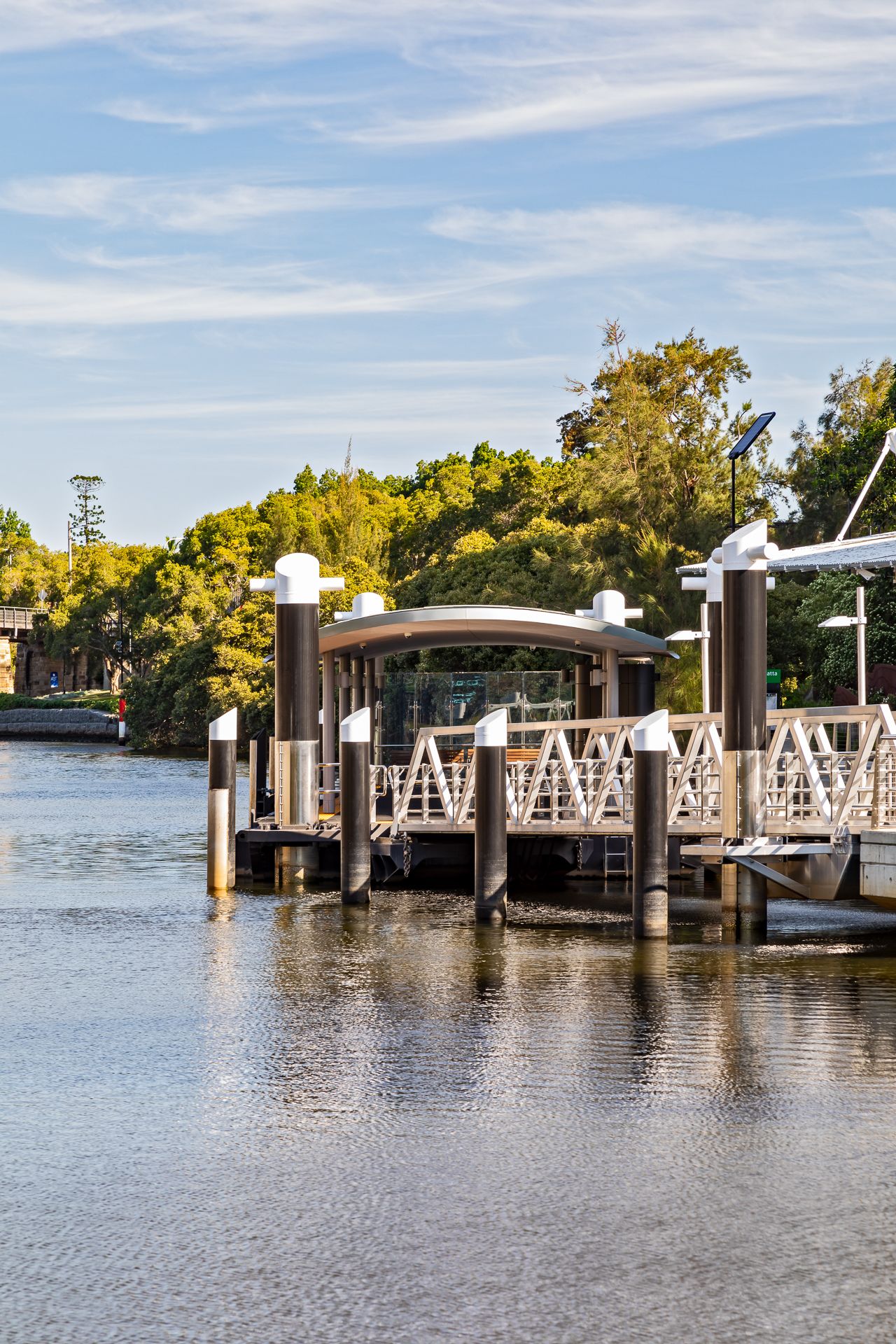 Modern ferry wharf with white mooring posts and a gangway on a still river, framed by green trees and soft clouds.
