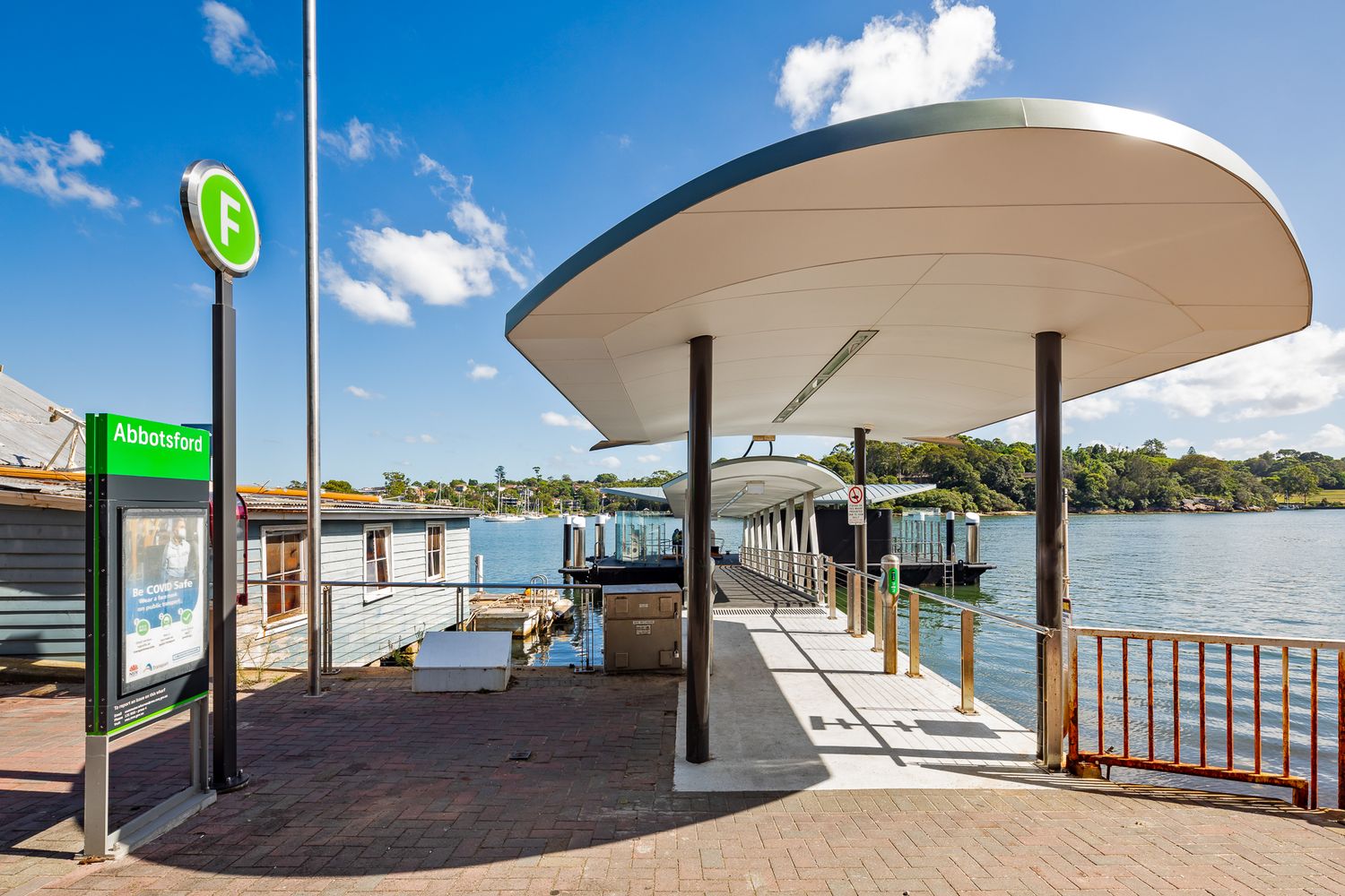 Ferry wharf entrance with green F ferry sign, curved canopy and timber railings beside harbour water and bushland on a clear day.