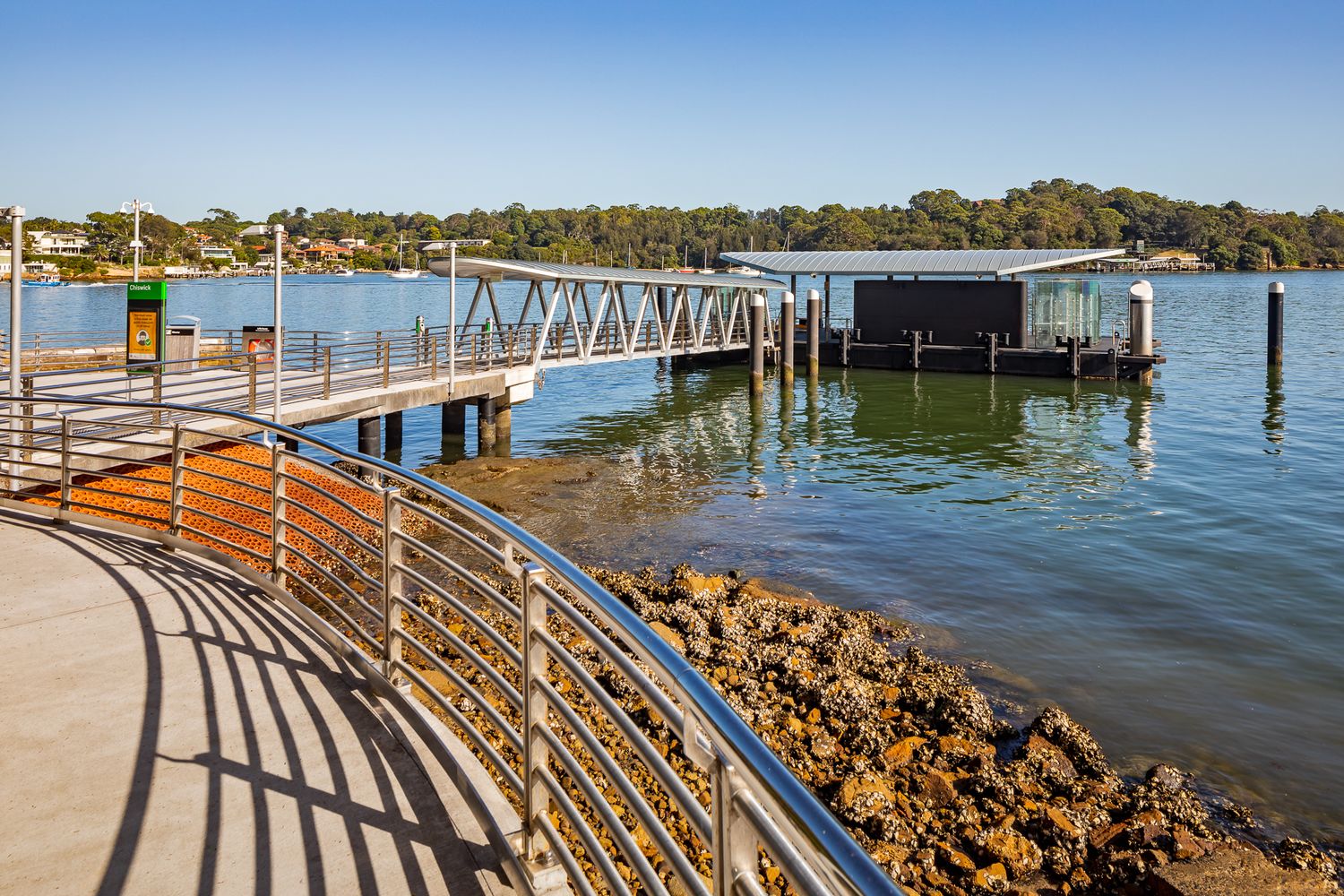 Long wooden jetty and ferry wharf extending over harbour waters, with a rocky shoreline and a curved metal railing in the foreground.