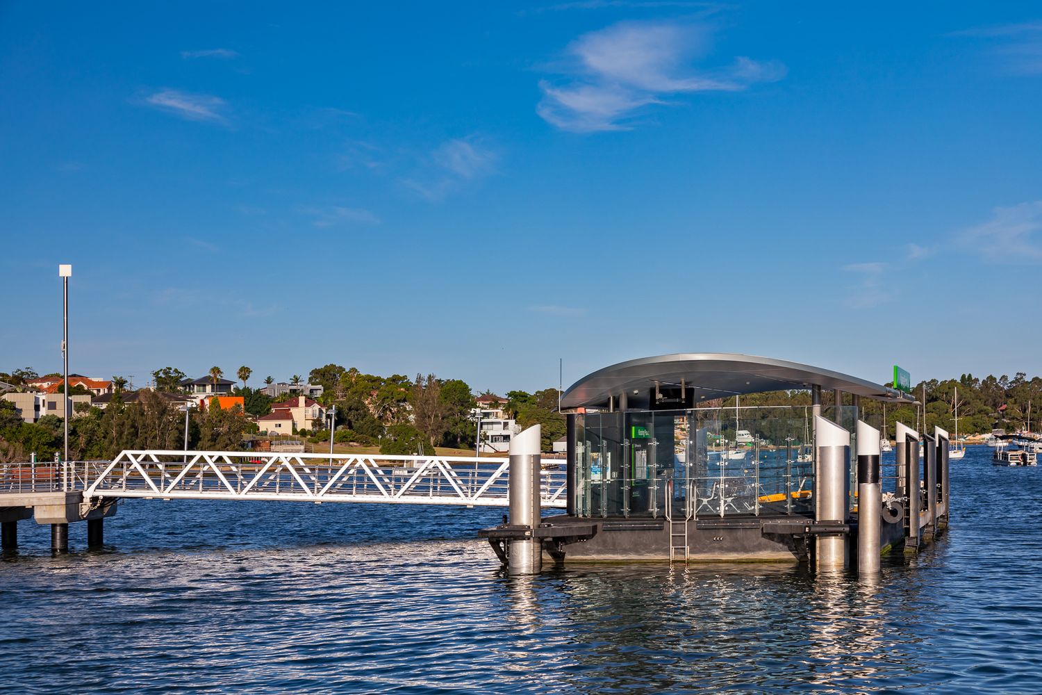 Glass ferry terminal with a curved roof and white gangway on blue harbour water, with waterfront homes and trees beyond.